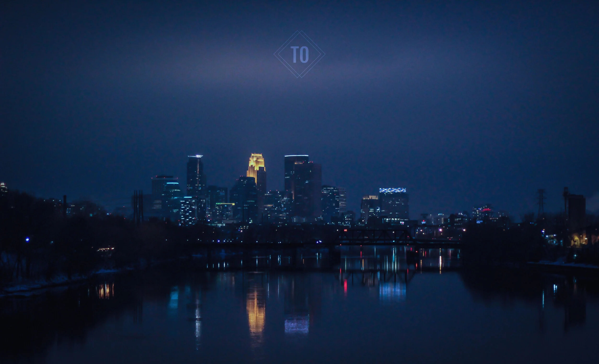 Nighttime city skyline with illuminated skyscrapers reflecting on a river in the foreground.