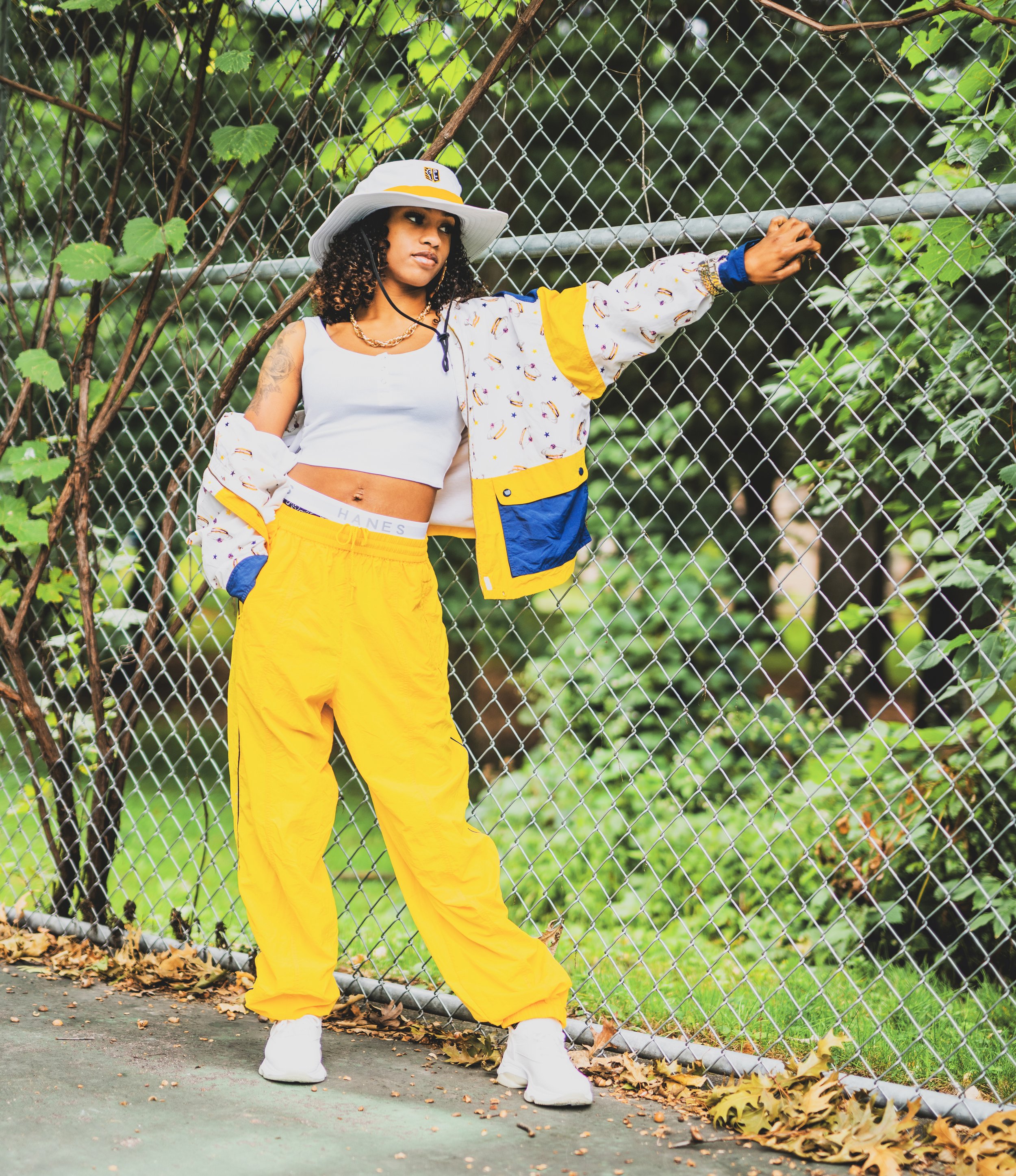 A young woman with curly hair standing next to a chain-link fence, wearing a white crop top, yellow pants, a colorful patterned jacket, a wide-brim hat, and white sneakers. She is outdoors with green foliage in the background.