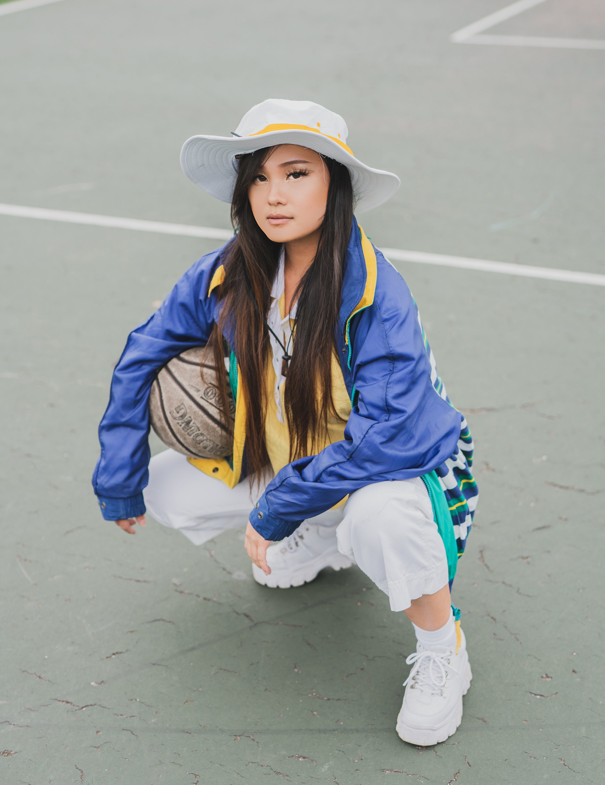 A woman squatting on a tennis court holding a basketball, wearing a large white hat, a blue jacket, a yellow shirt, white pants, and white sneakers.