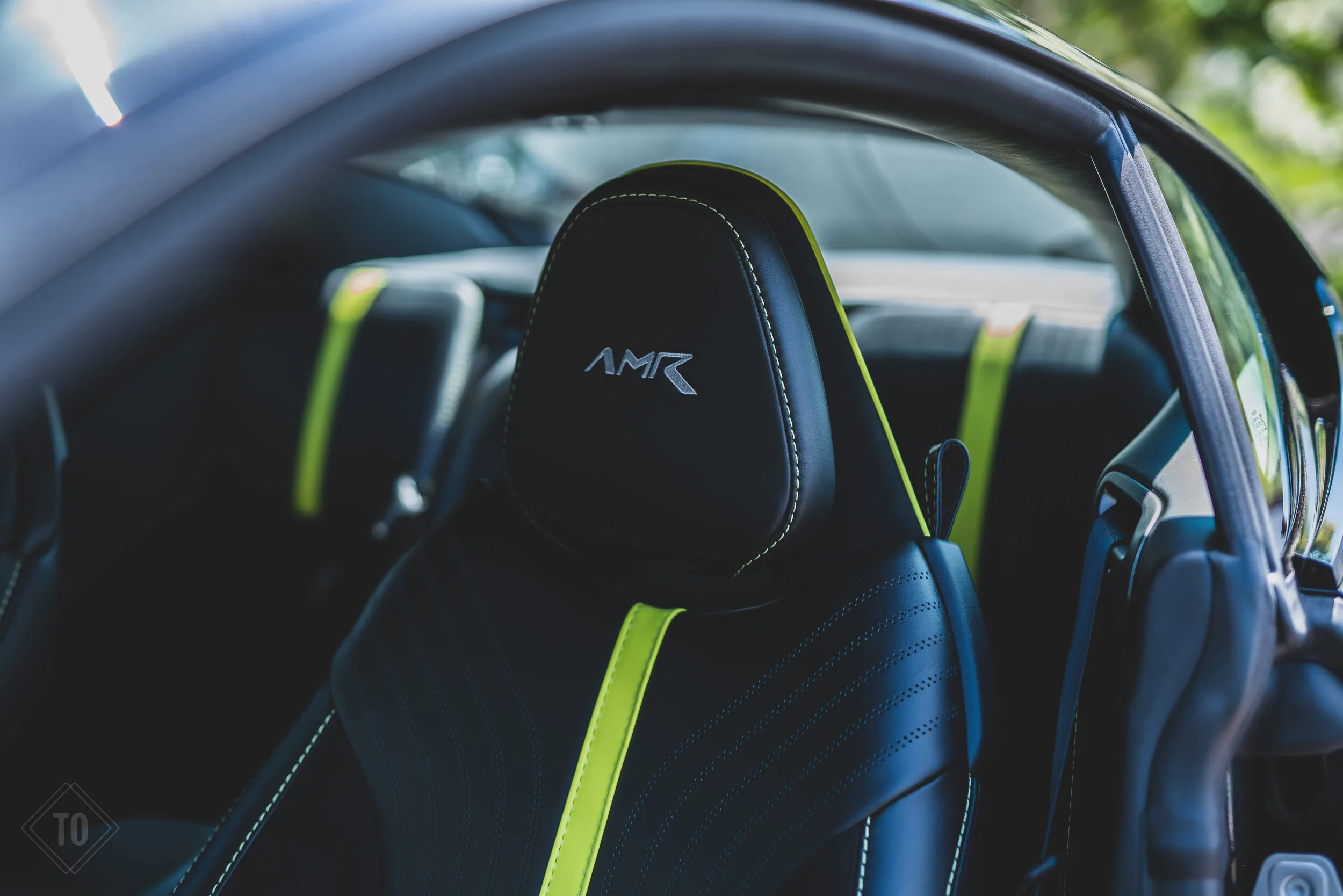 Close-up of a black car seat with white stitching and yellow-green accents, featuring 'AMR' embroidered on the headrest.