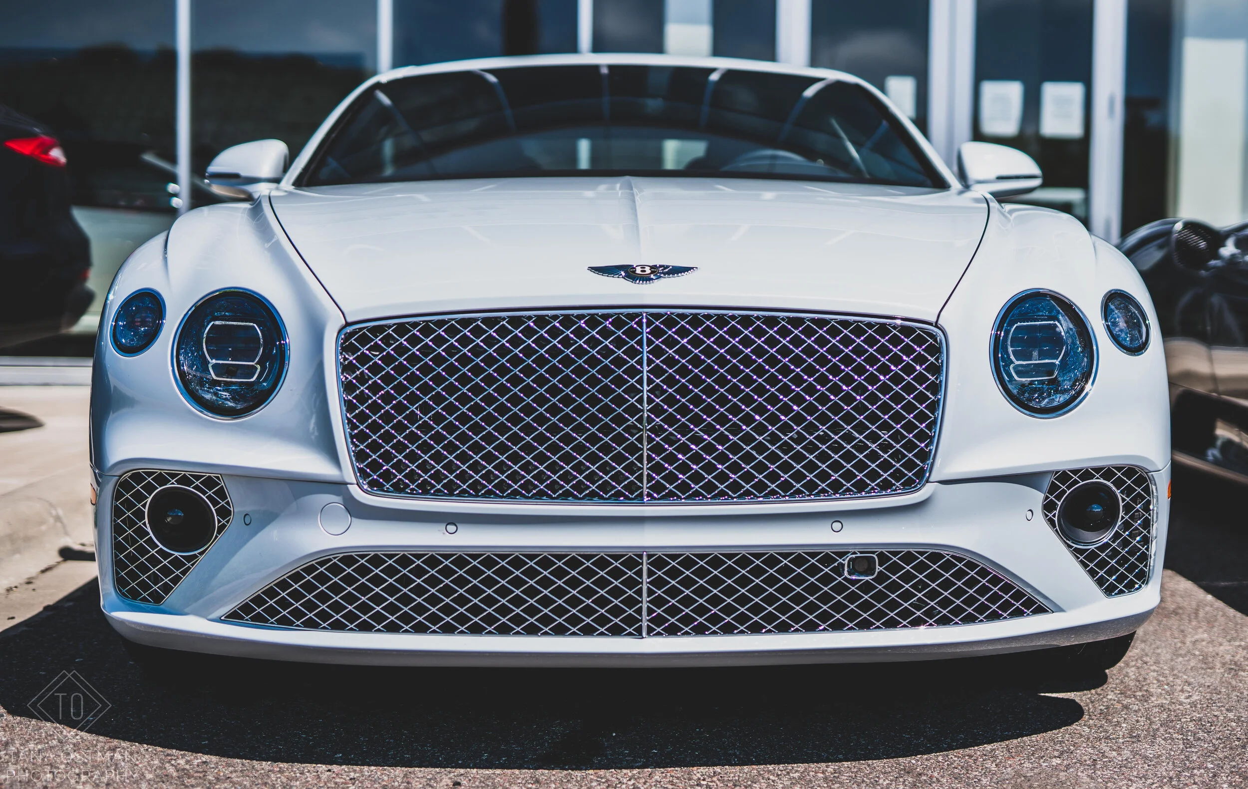 Front view of a white luxury car with a large grille, circular headlights, and a Bentley logo on the hood, parked outside a dealership.
