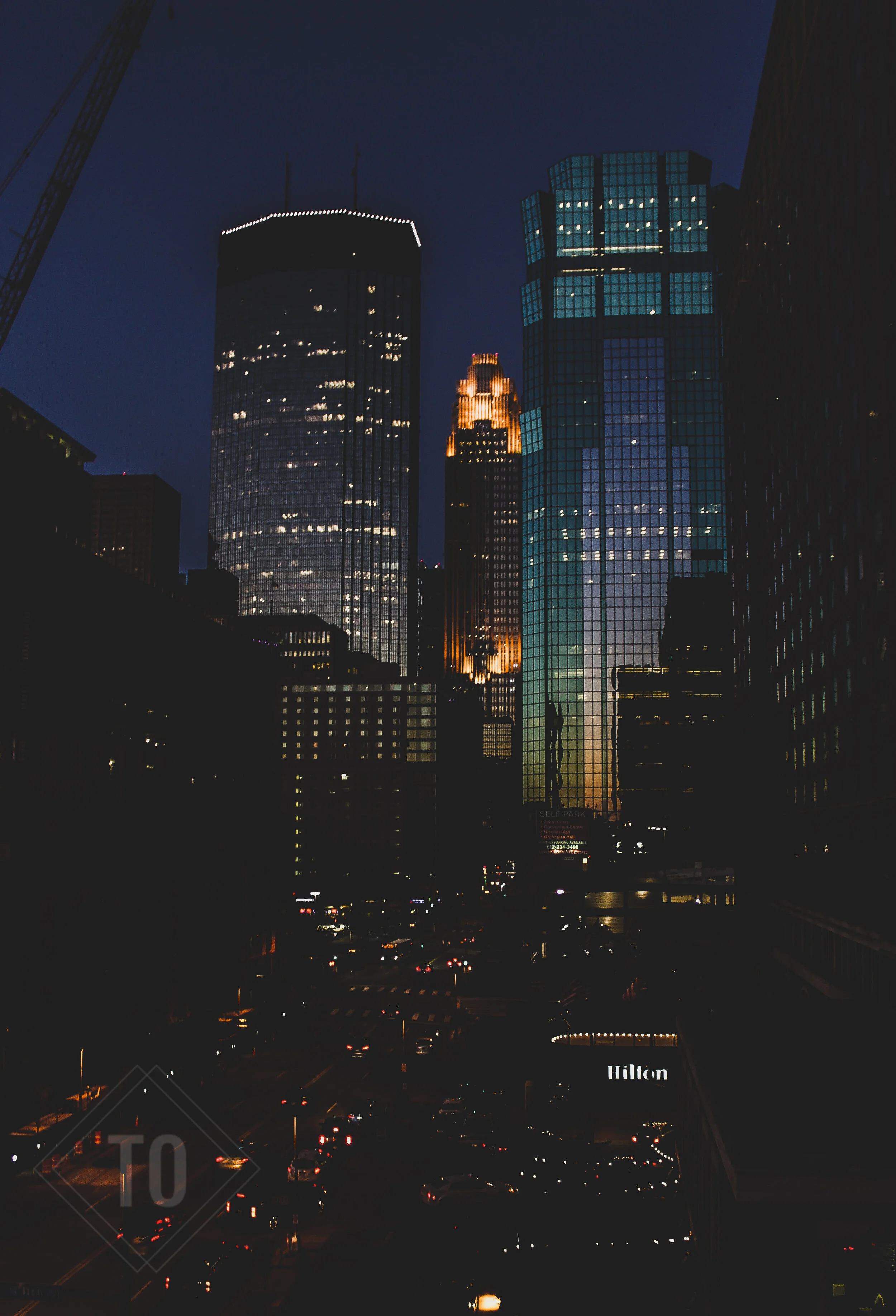 Night view of a city skyline with illuminated skyscrapers, including the Hilton hotel, and traffic on the street below.