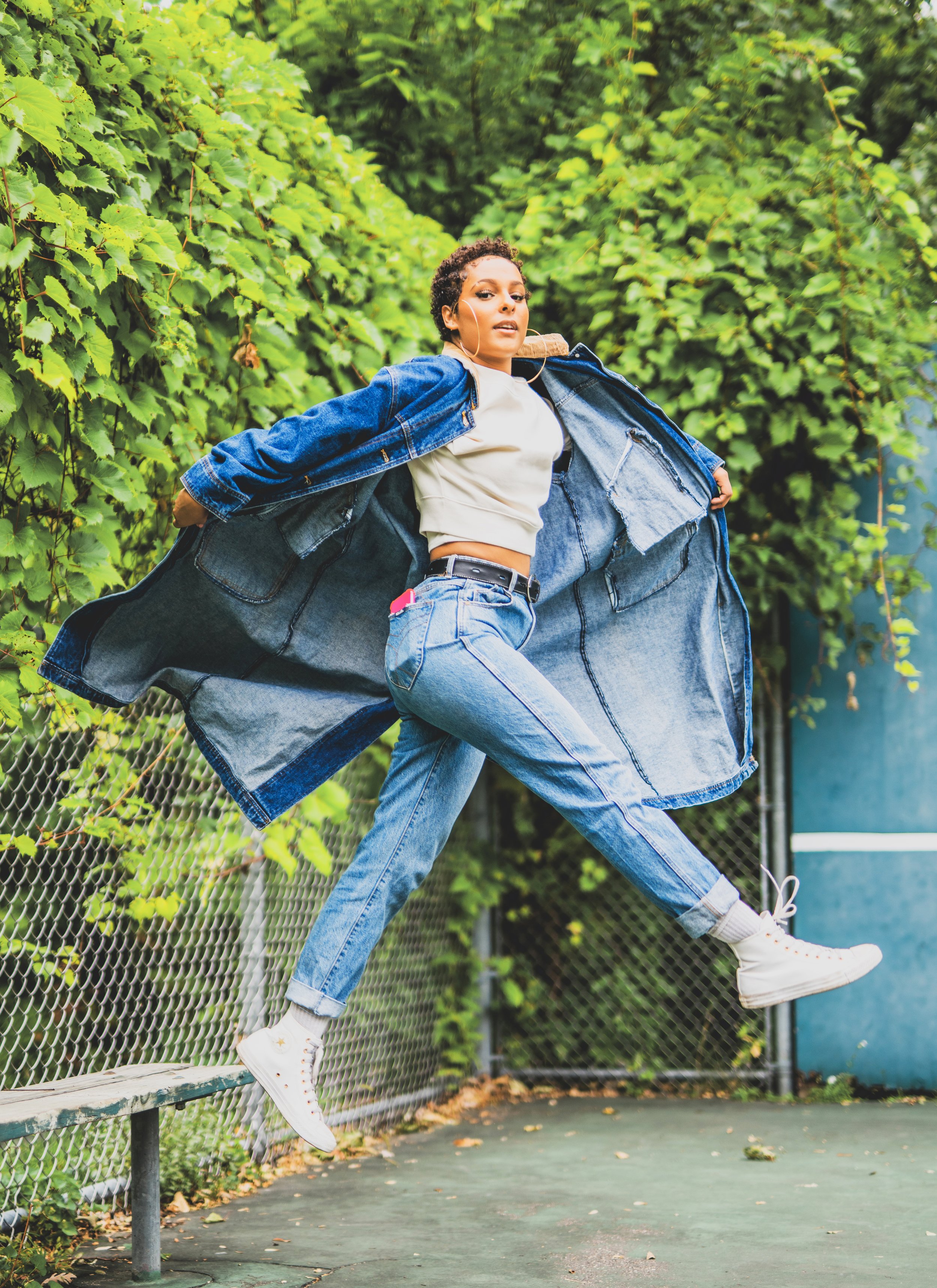 A young woman in a denim jacket, white crop top, and jeans is levitating or jumping in front of green bushes and a chain-link fence, with a sporty environment.