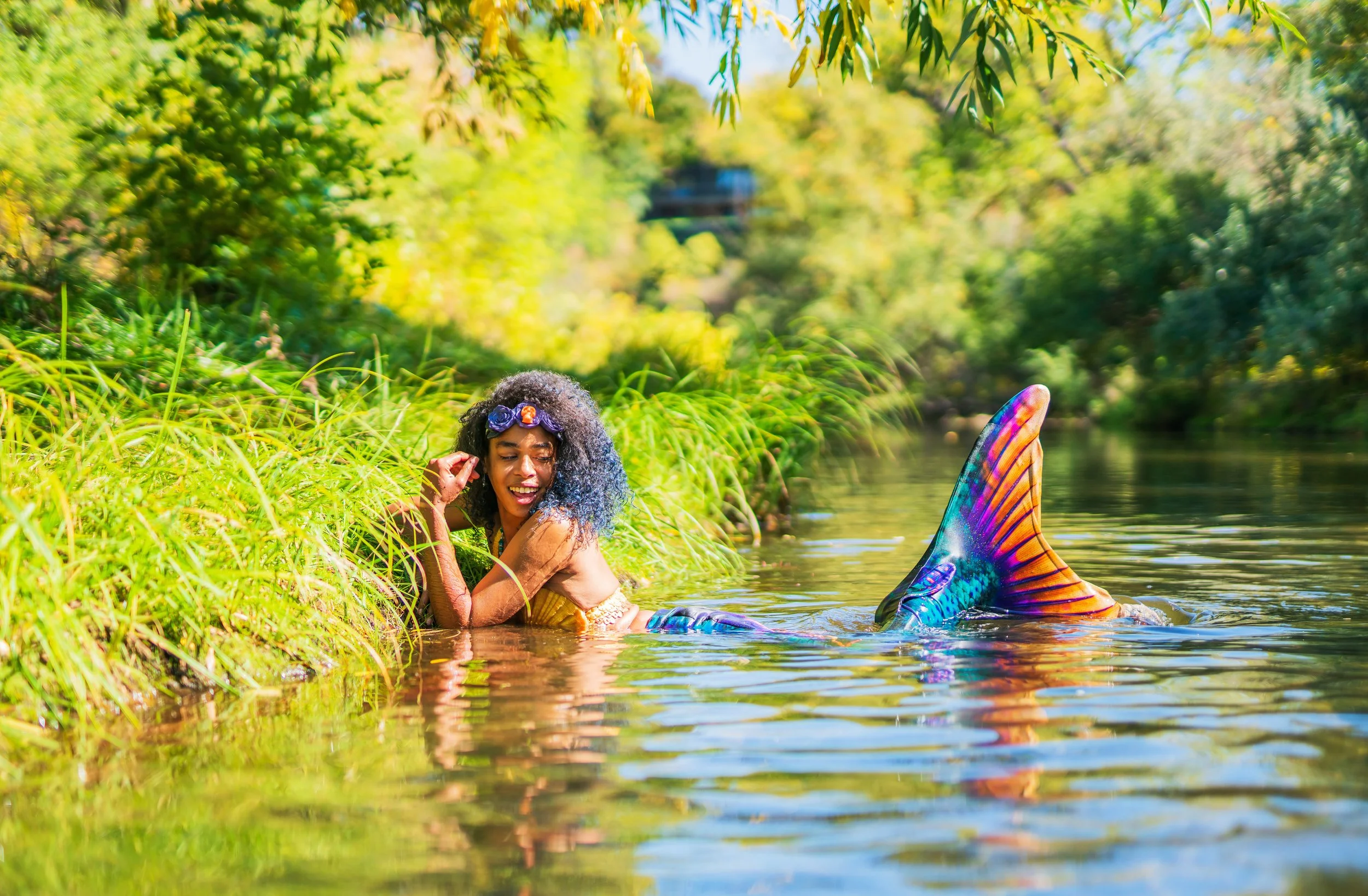 A woman dressed as a mermaid with a colorful tail, sitting in a river surrounded by green foliage and trees, smiling and enjoying the water.