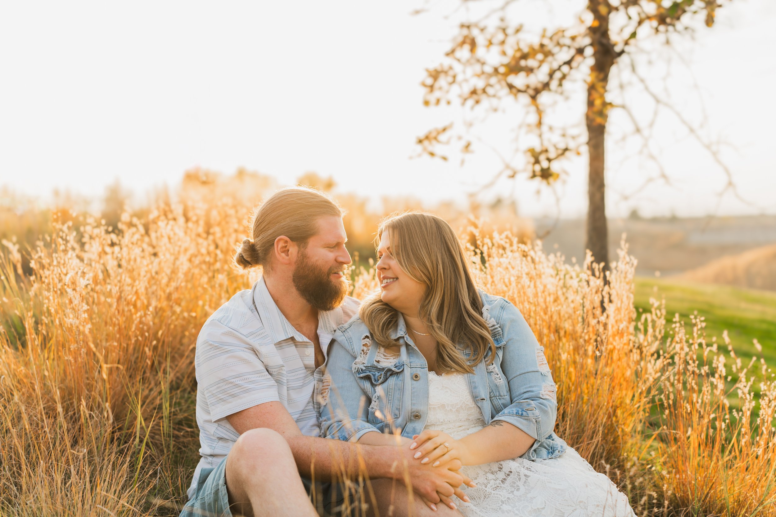 A couple sitting on the grass in a field of tall grass with a tree in the background, enjoying a moment together during sunset.