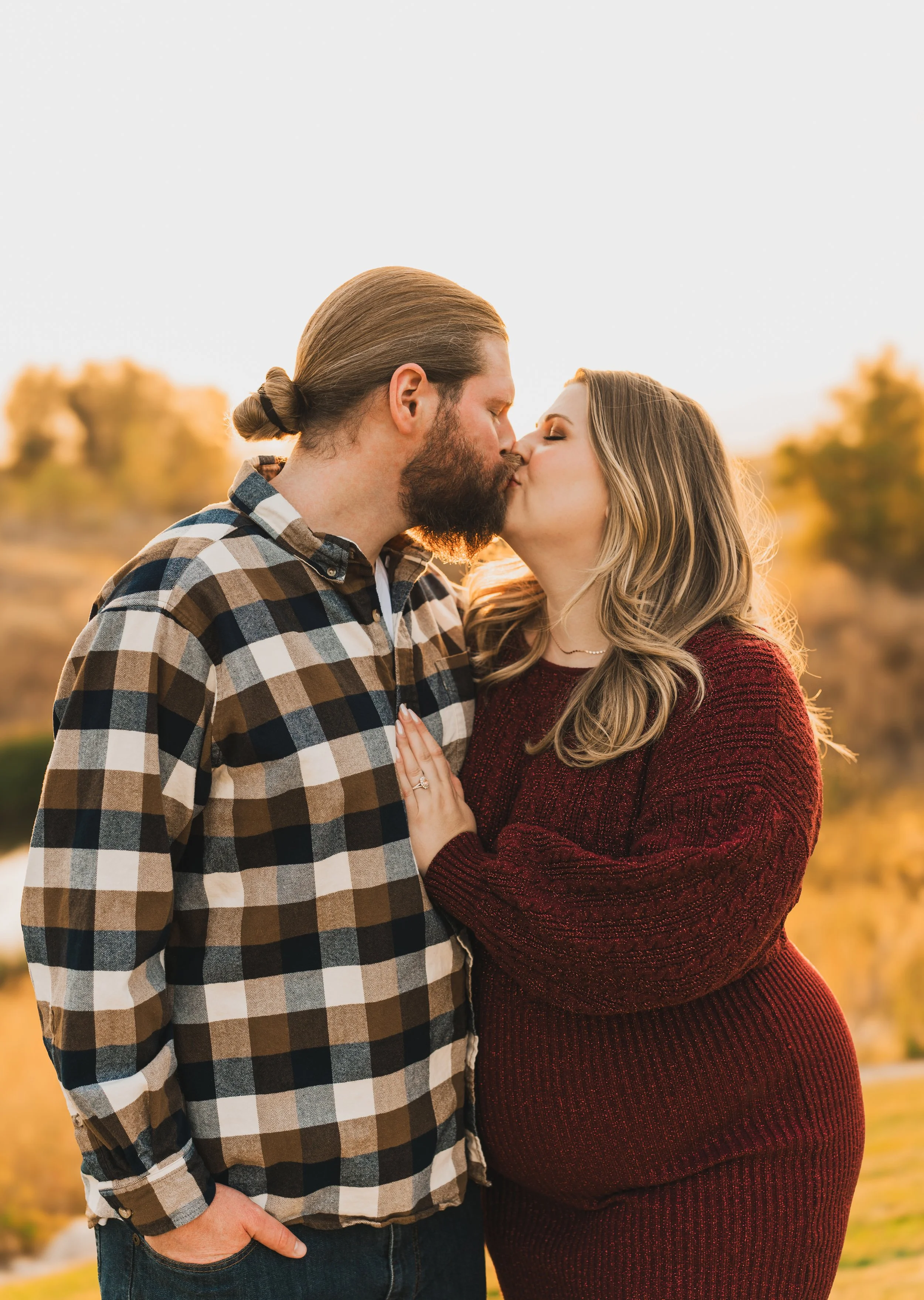 A couple kissing outdoors during sunset, with autumn trees in the background.