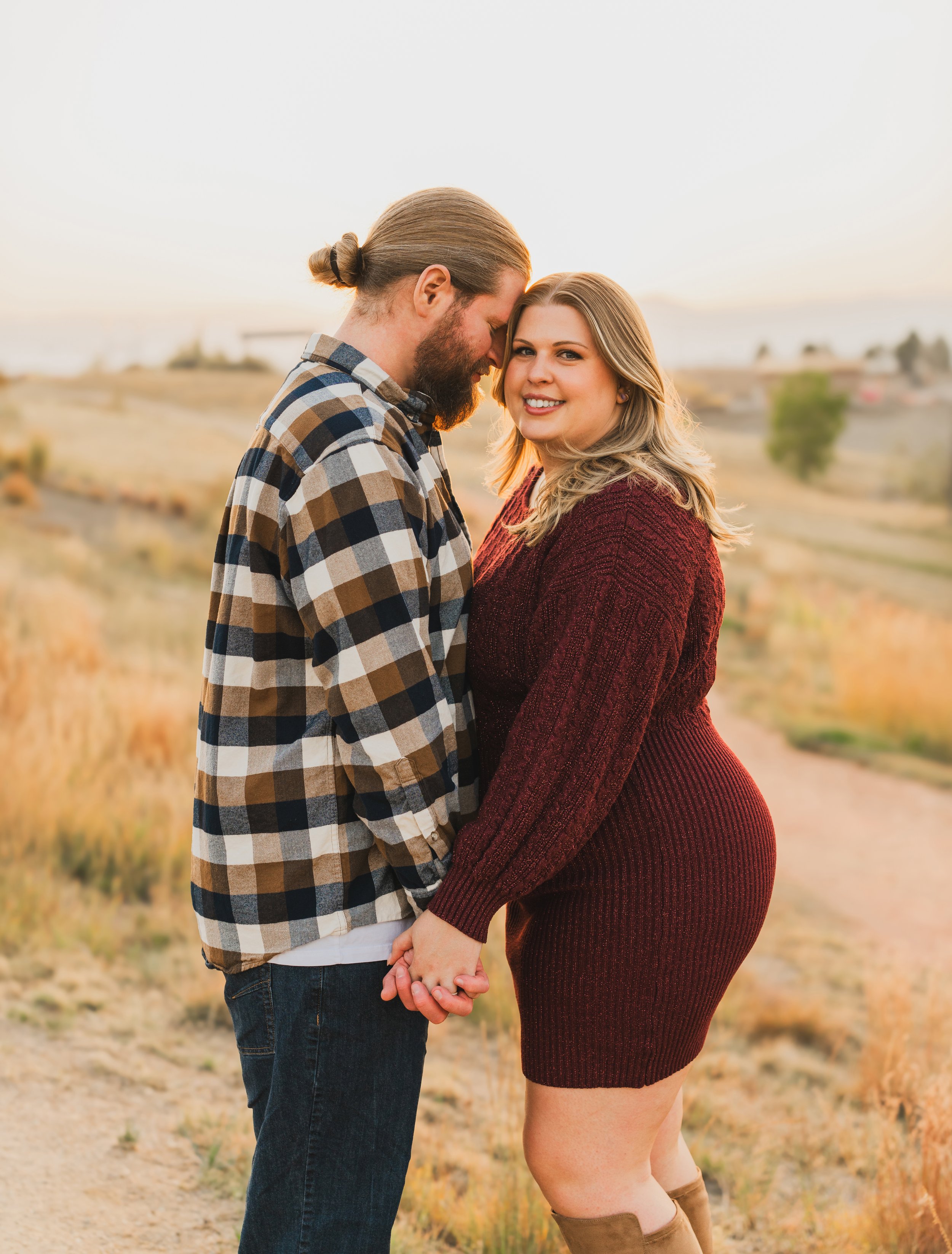 A couple holding hands and smiling outdoors during sunset, with a dirt path and grassy field in the background.