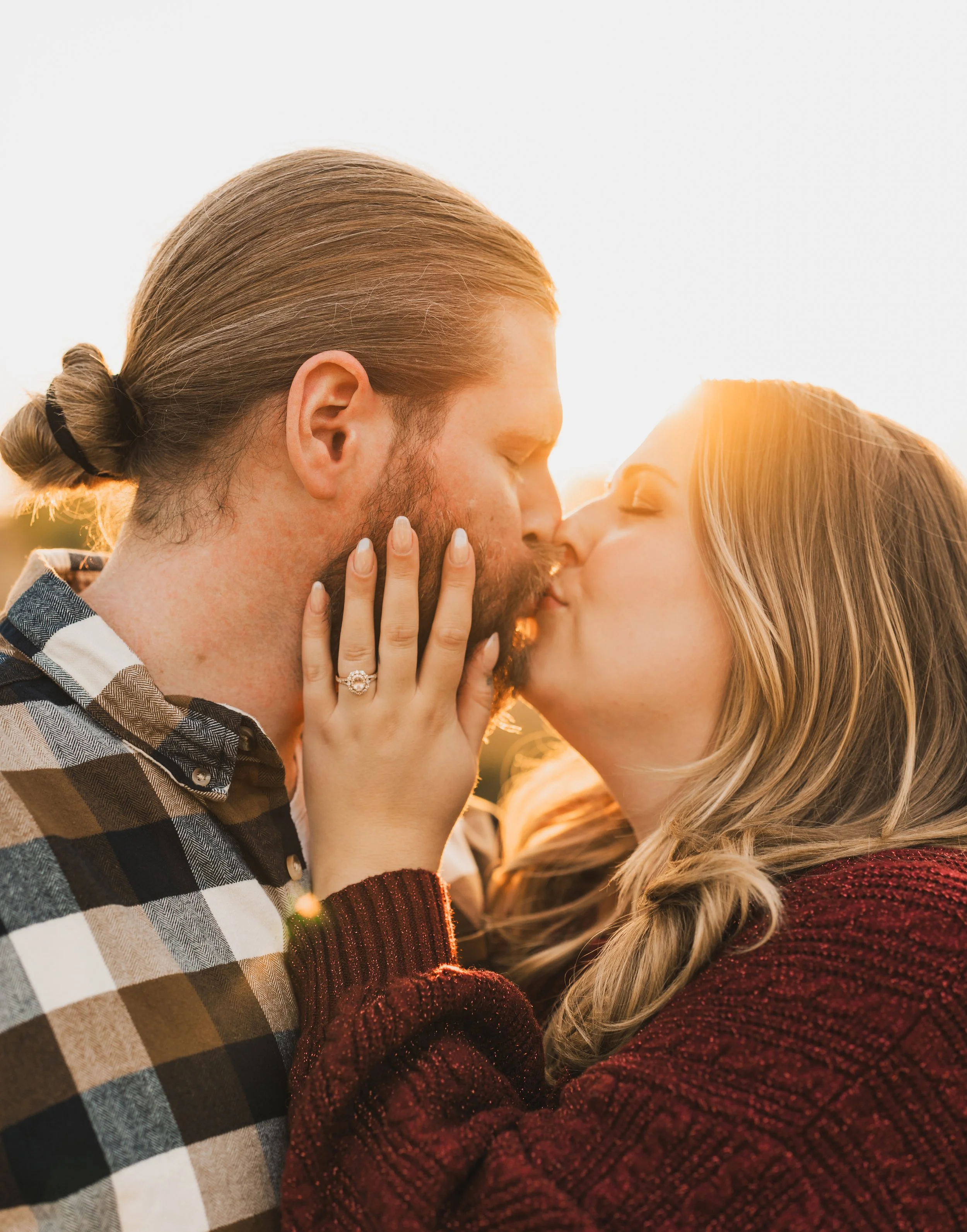 A couple kissing outdoors at sunset, the woman with blonde hair and the man with a beard and tied-back hair, the woman showing her engagement ring.