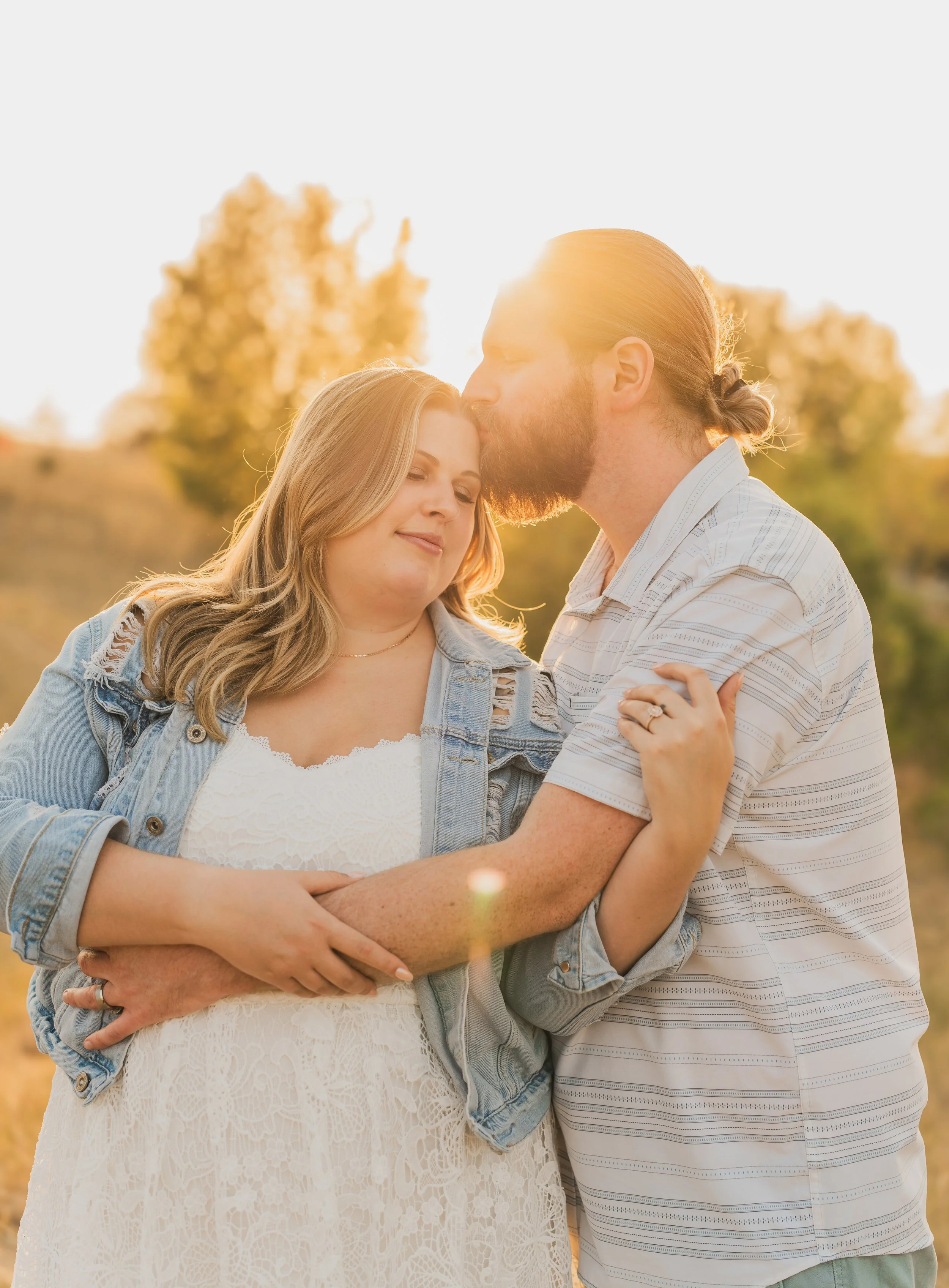 A couple embracing outdoors during sunset, with the man kissing the woman's forehead. The woman has blonde hair and is wearing a white lace dress with a ripped denim jacket. The man has reddish hair, a beard, and is wearing a striped shirt. They are 