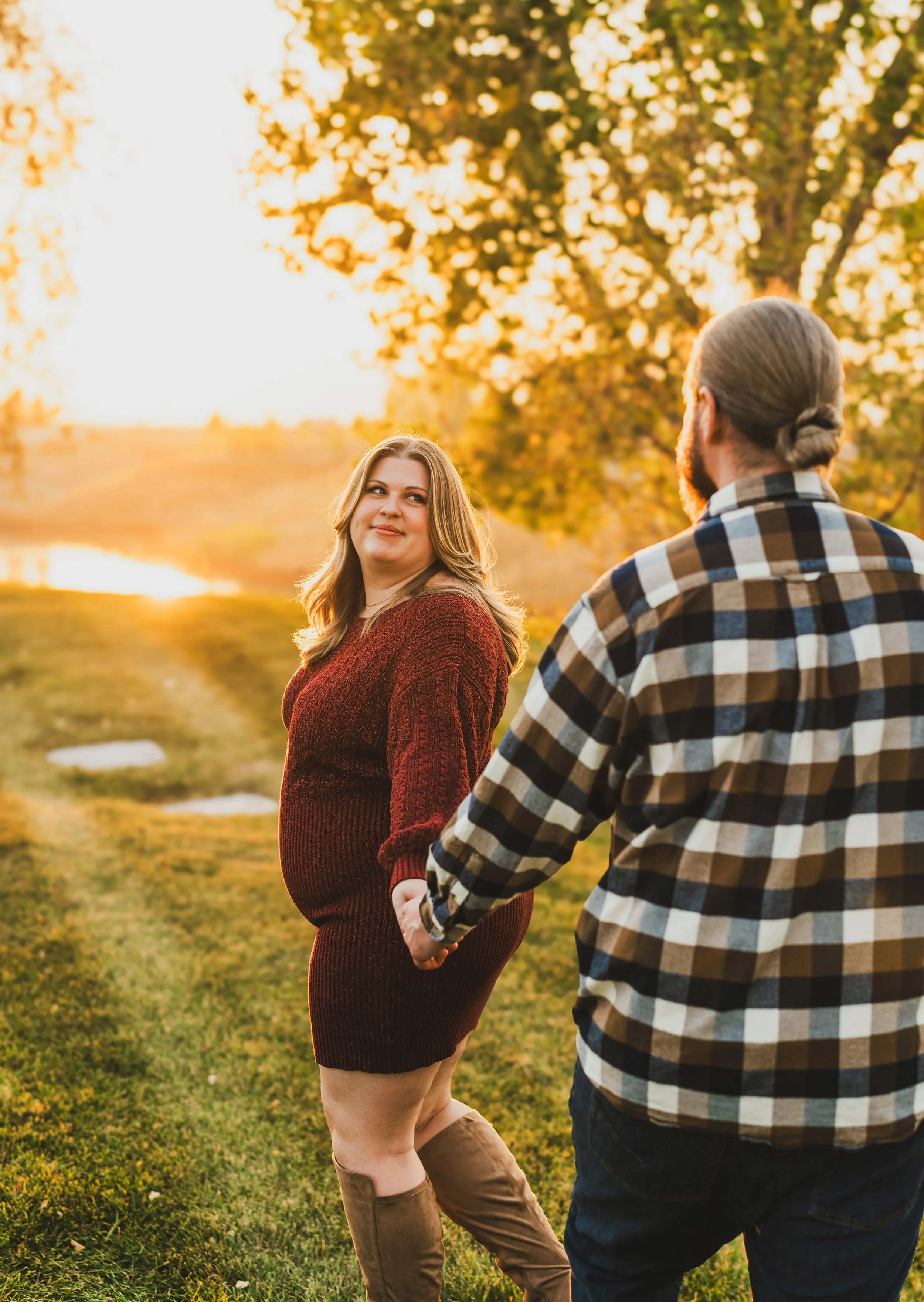 A couple holding hands and walking outdoors during sunset, with trees and a pond in the background, autumn foliage, and warm clothing.