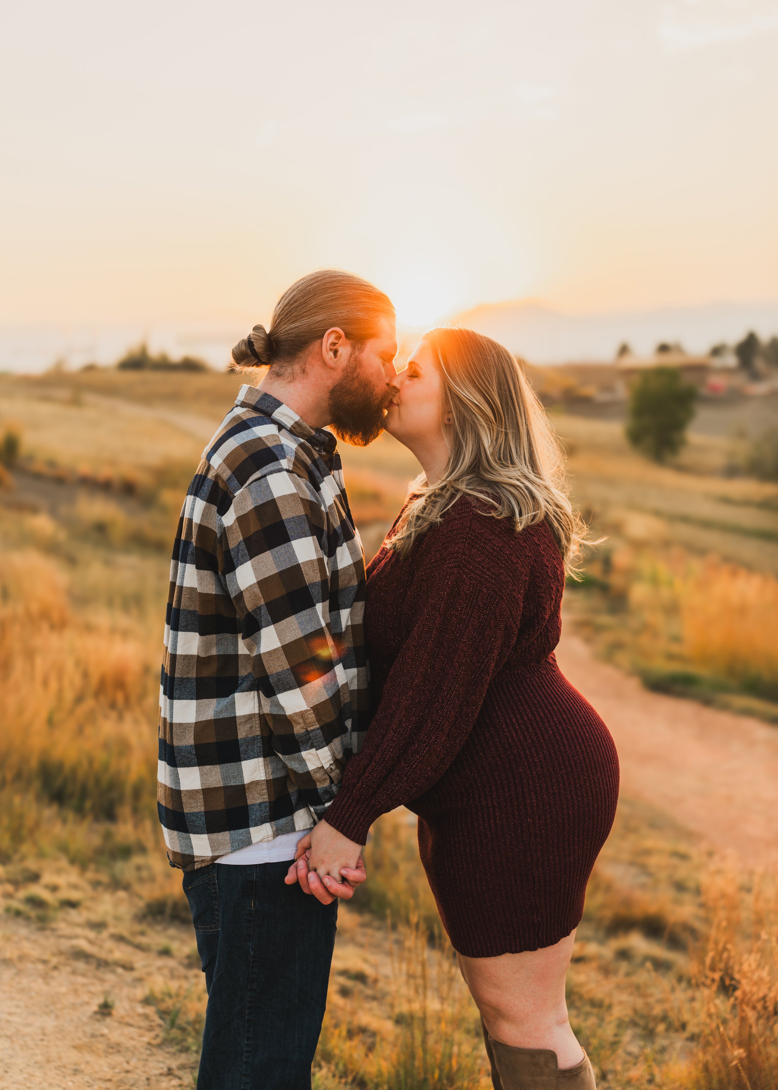 A couple kissing outdoors during sunset in a field, holding hands, with a rural landscape in the background.