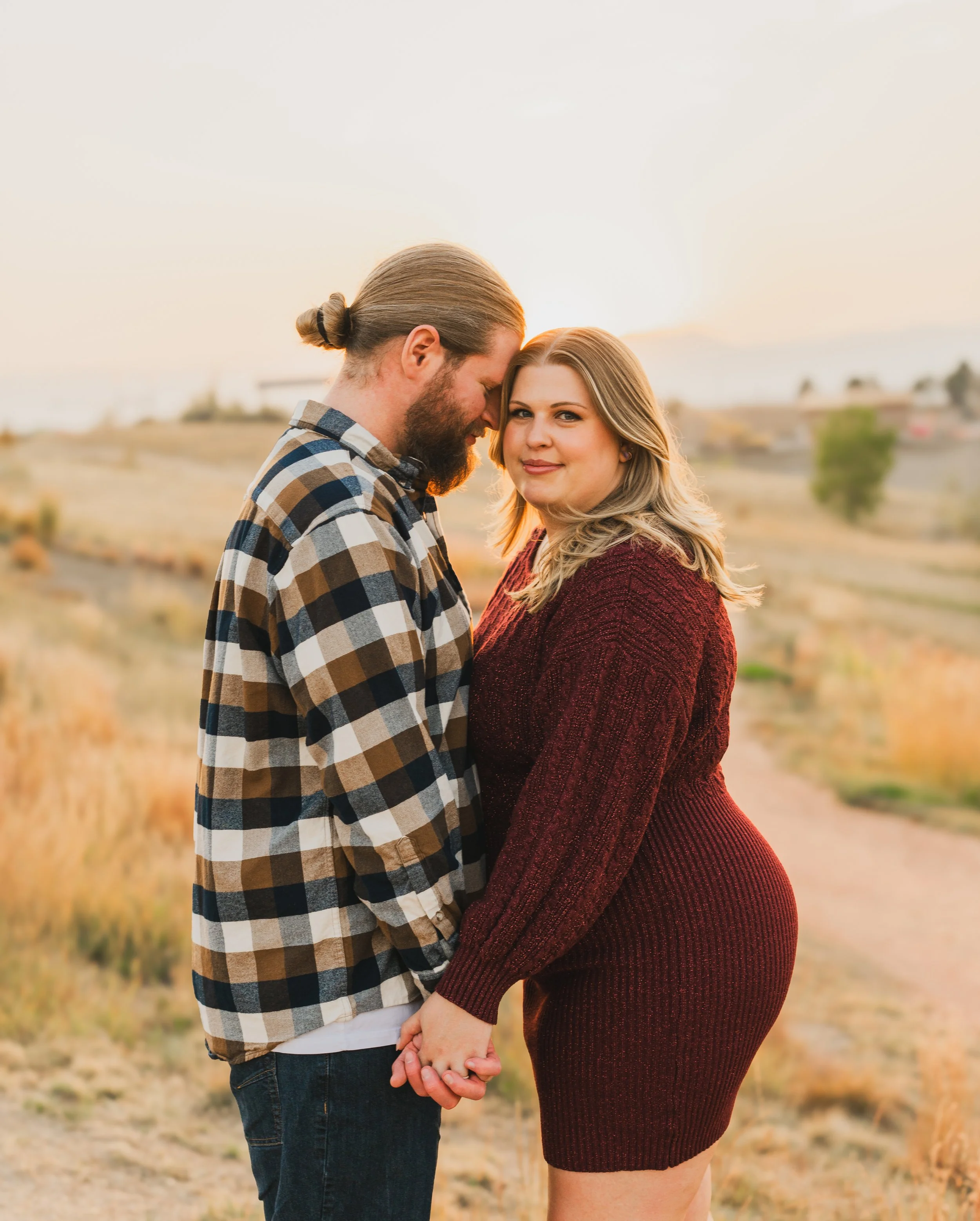 A couple holding hands outdoors at sunset, standing on a dirt path with a rural landscape in the background.