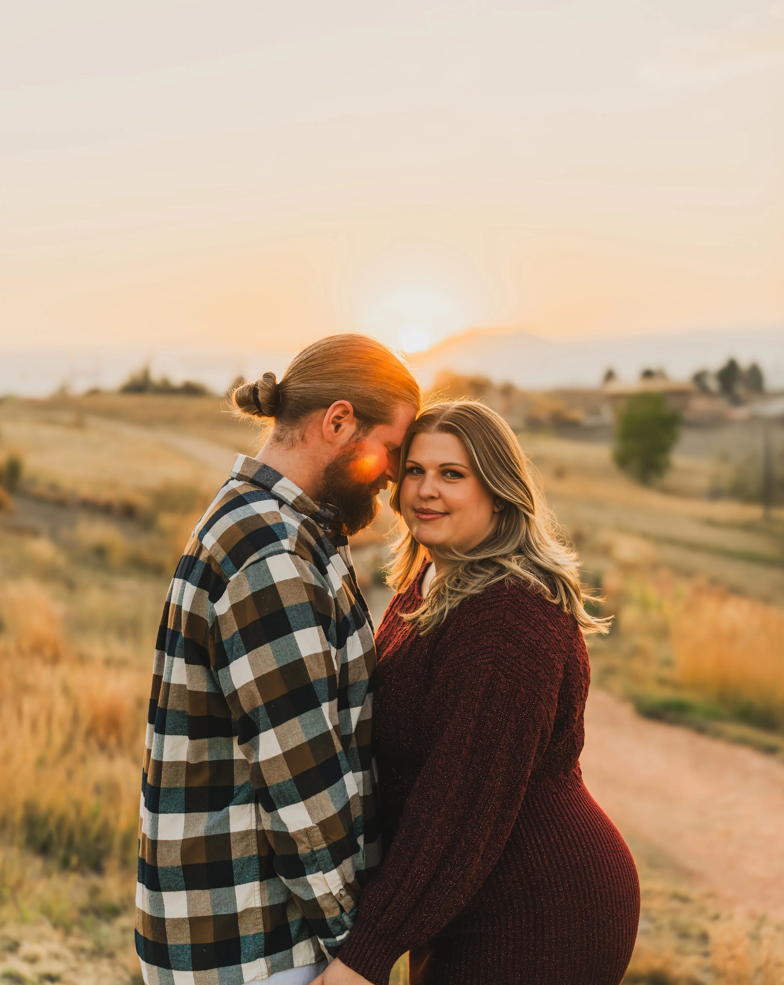 A couple standing close together outdoors during sunset, with rolling hills and a dirt path in the background.