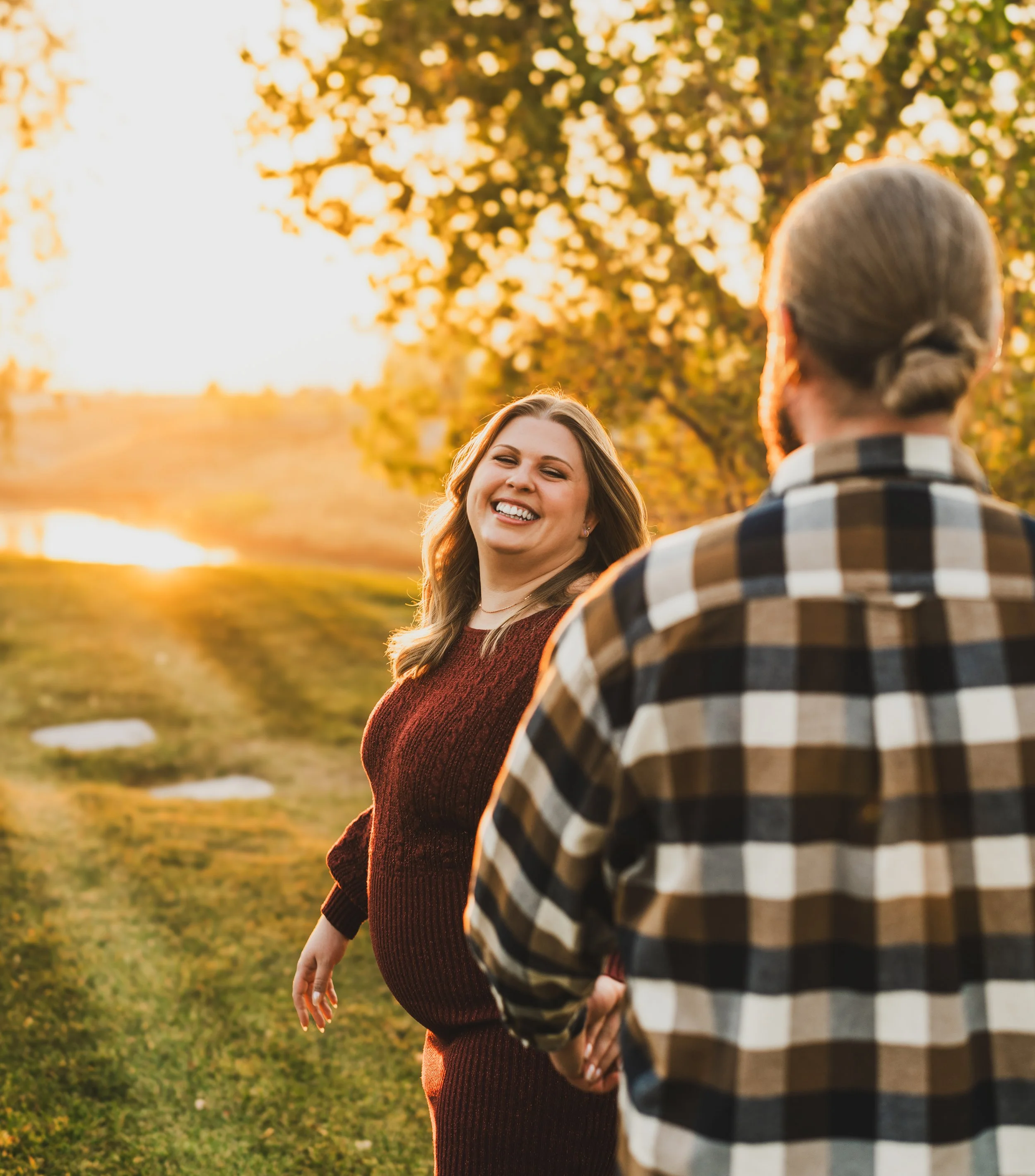 A smiling pregnant woman in a maroon sweater standing outdoors during sunset, talking with a man in a plaid shirt with long hair tied back in a ponytail, in a park with trees and a lake in the background.