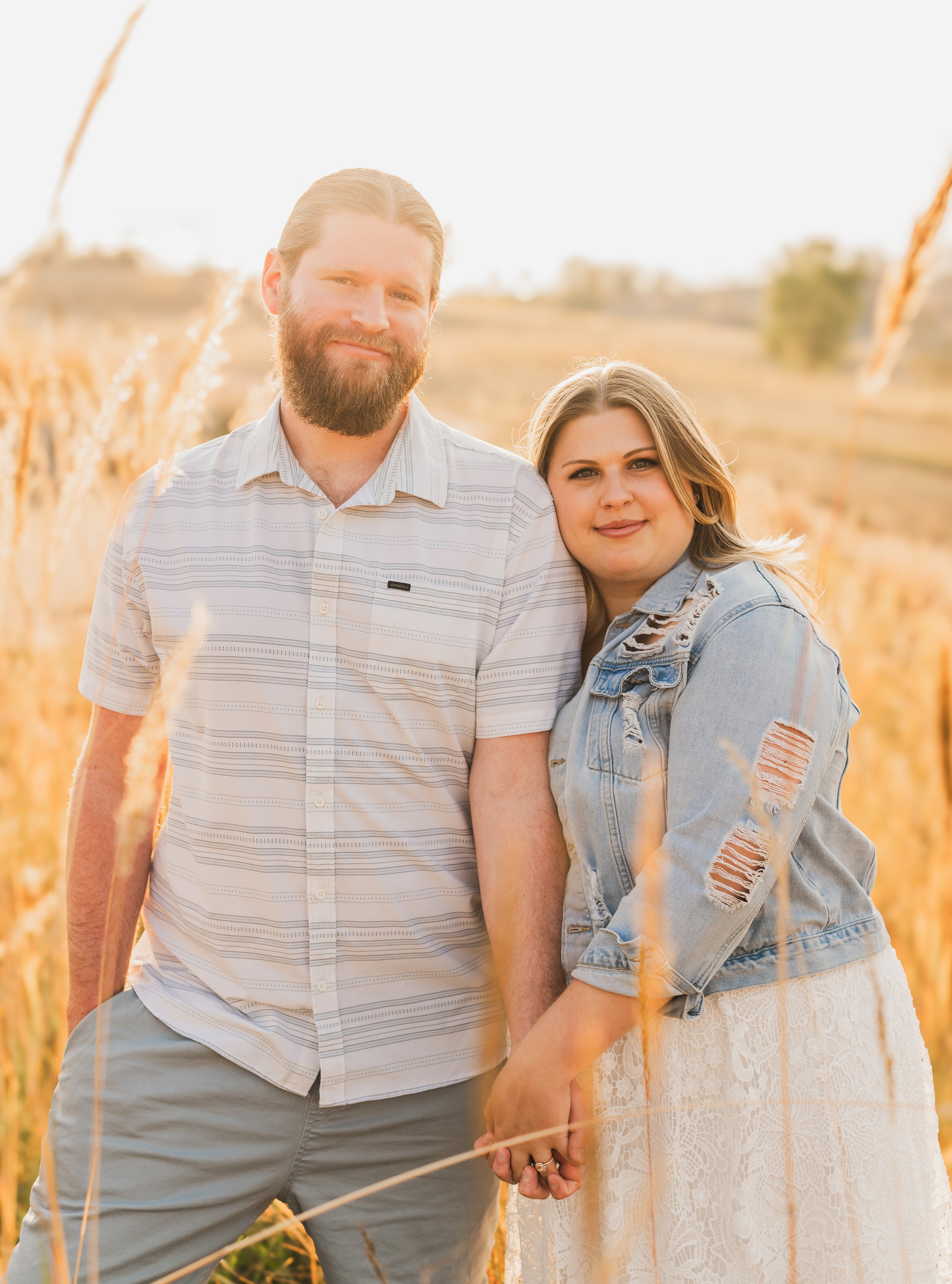 A young couple holding hands in a field of tall, golden grass during sunset, with a blurred natural landscape in the background.