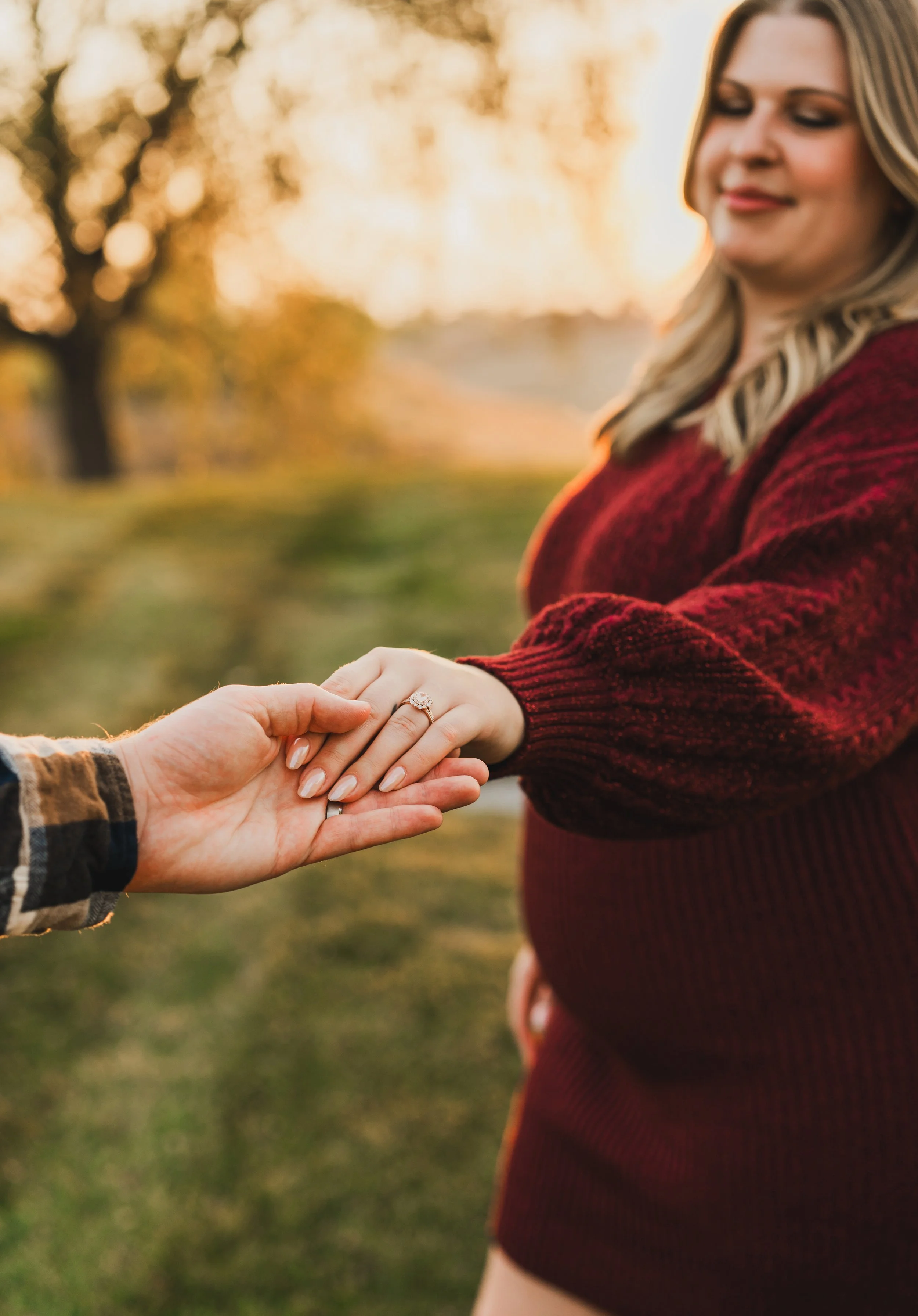 A pregnant woman in a red sweater holding hands with another person outdoors during sunset.