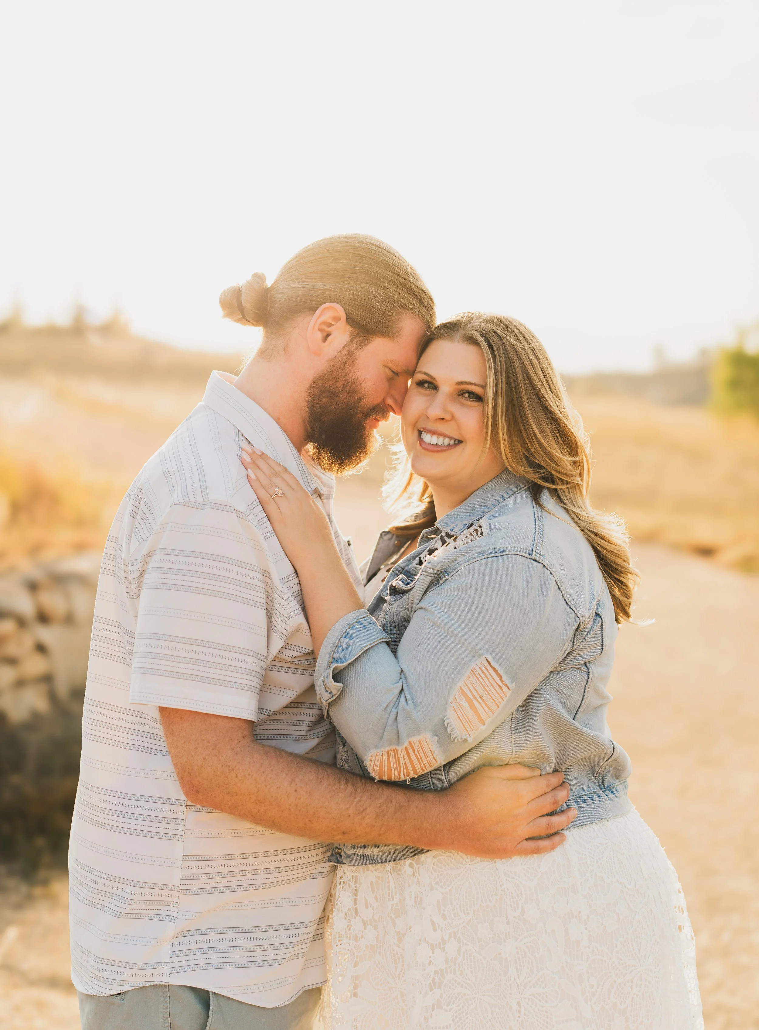 A smiling woman and a man with long hair and beard embrace outdoors in a sunny, rural landscape.