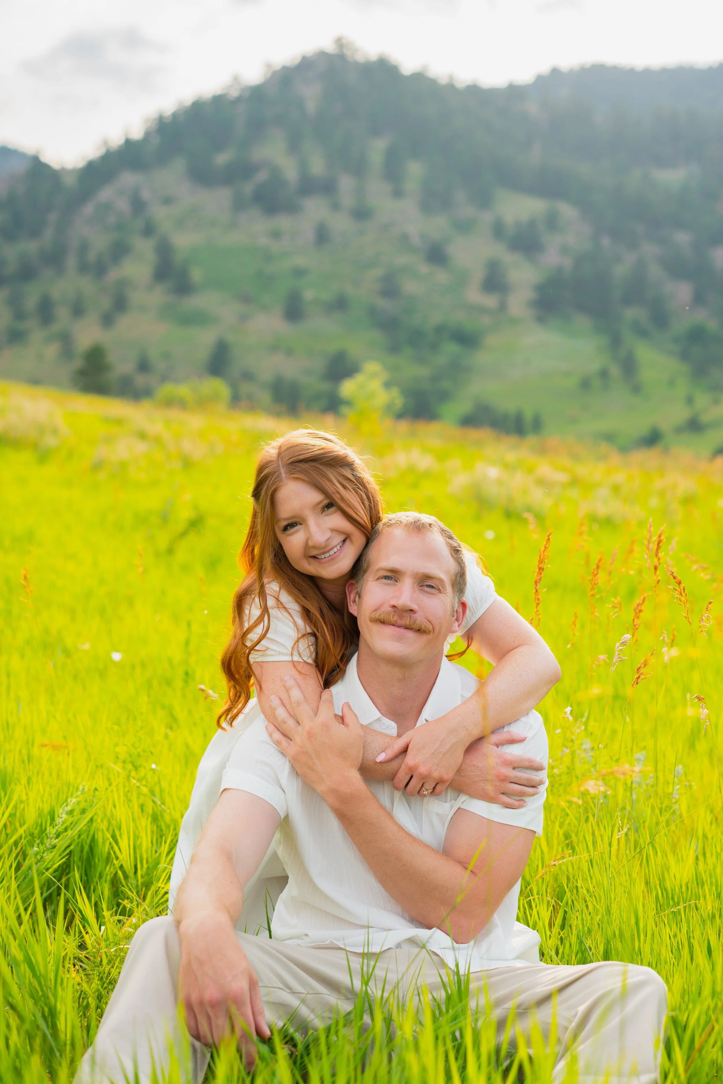 A happy couple sitting and smiling in a lush green meadow with mountains in the background on a sunny day.