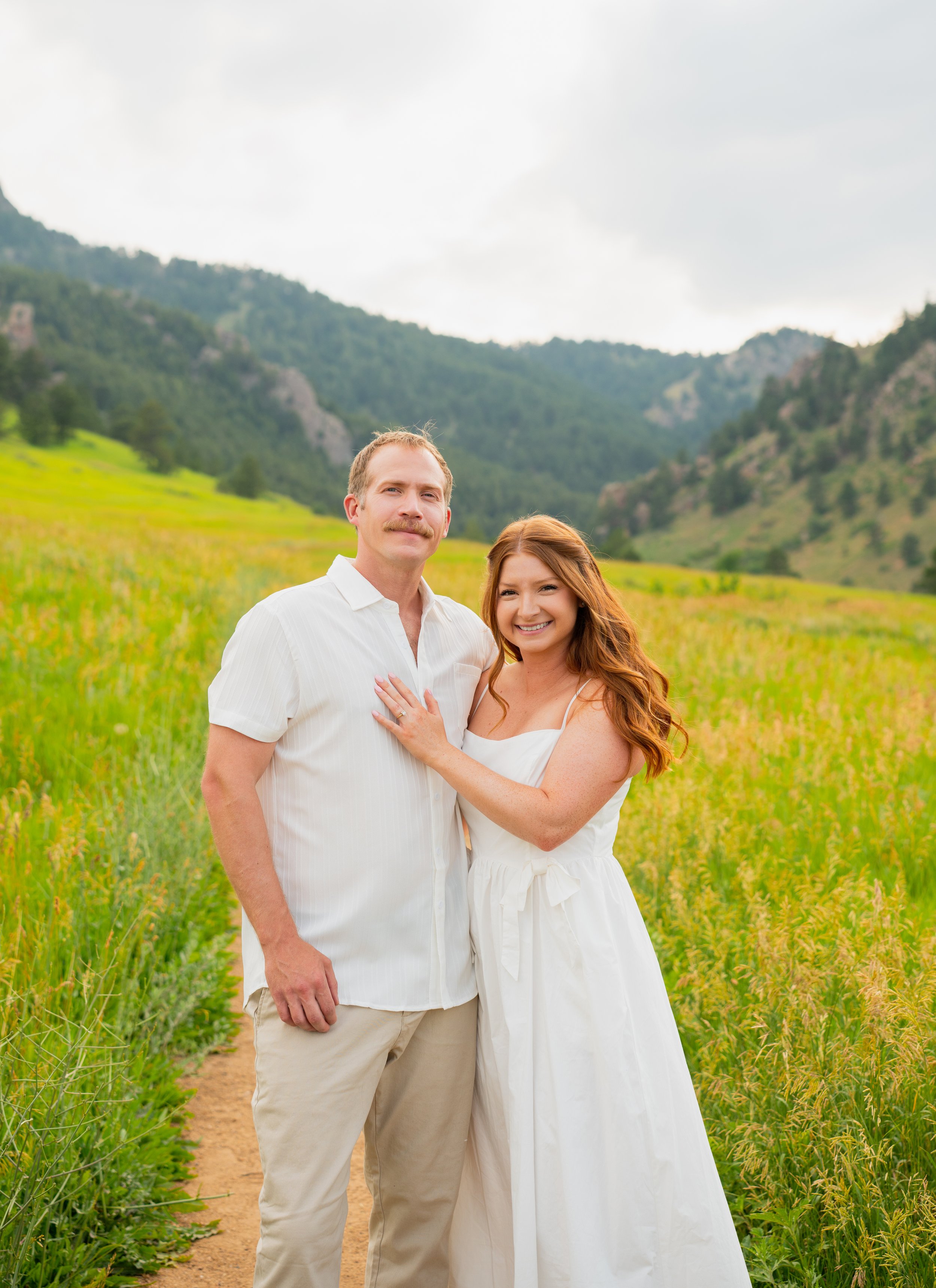 A happy couple stands arm in arm in a lush green meadow with mountains in the background, both dressed in white, smiling at the camera.