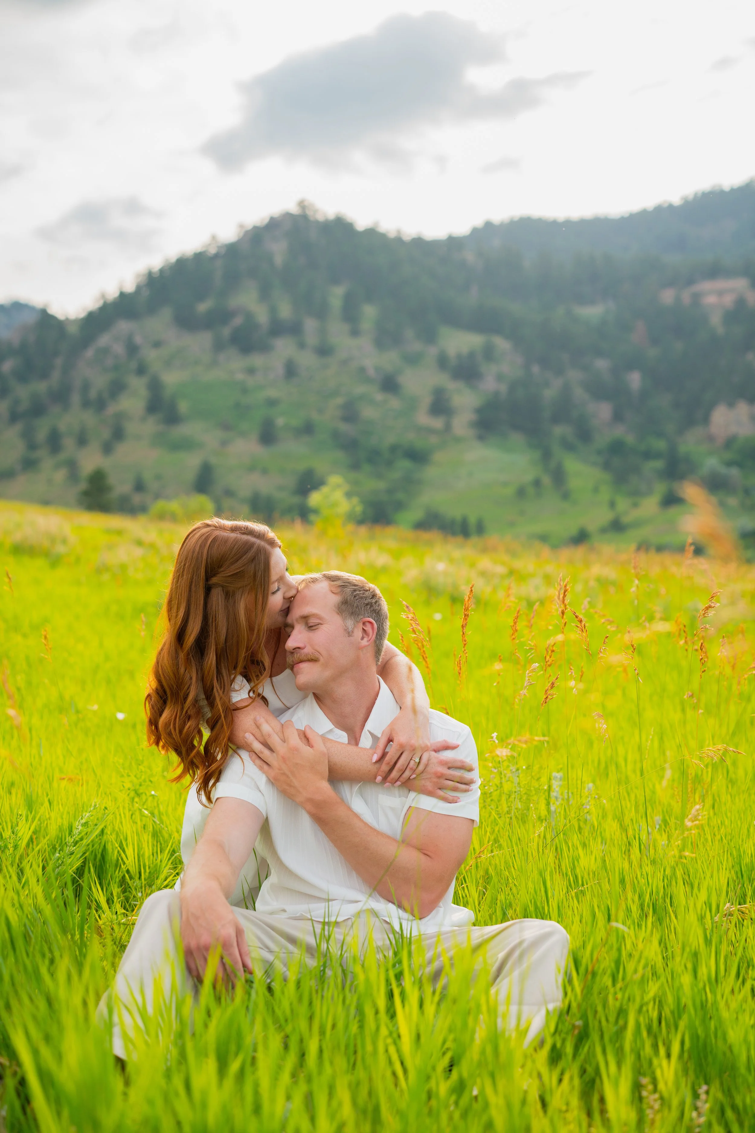 A couple sitting in a grassy field with mountains in the background. The woman is kissing the man's forehead while hugging him, both dressed in white.