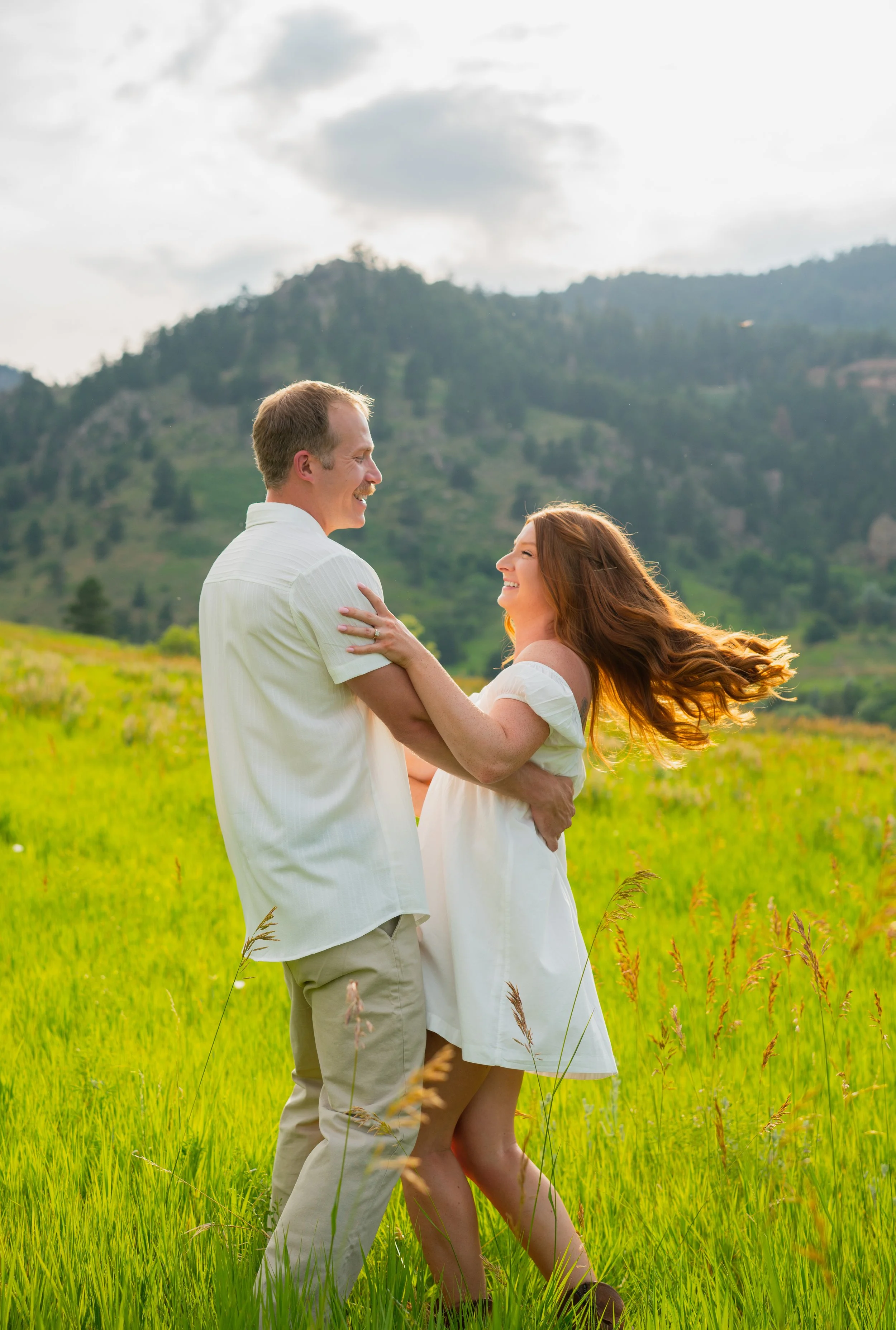 A happy couple is embracing in a lush green meadow with mountains in the background, under a cloudy sky.