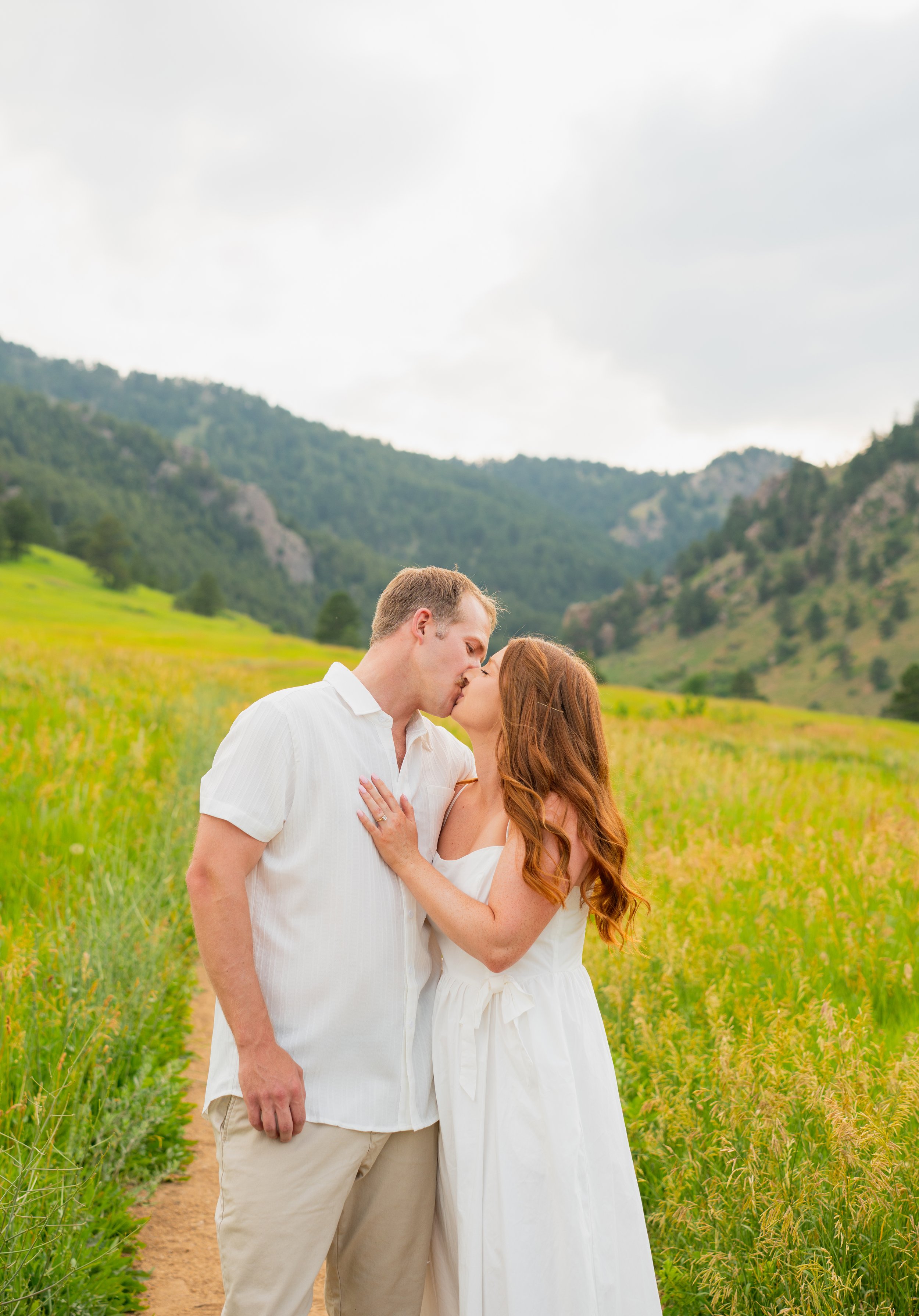 A couple kissing in a field with mountains in the background, dressed in white.