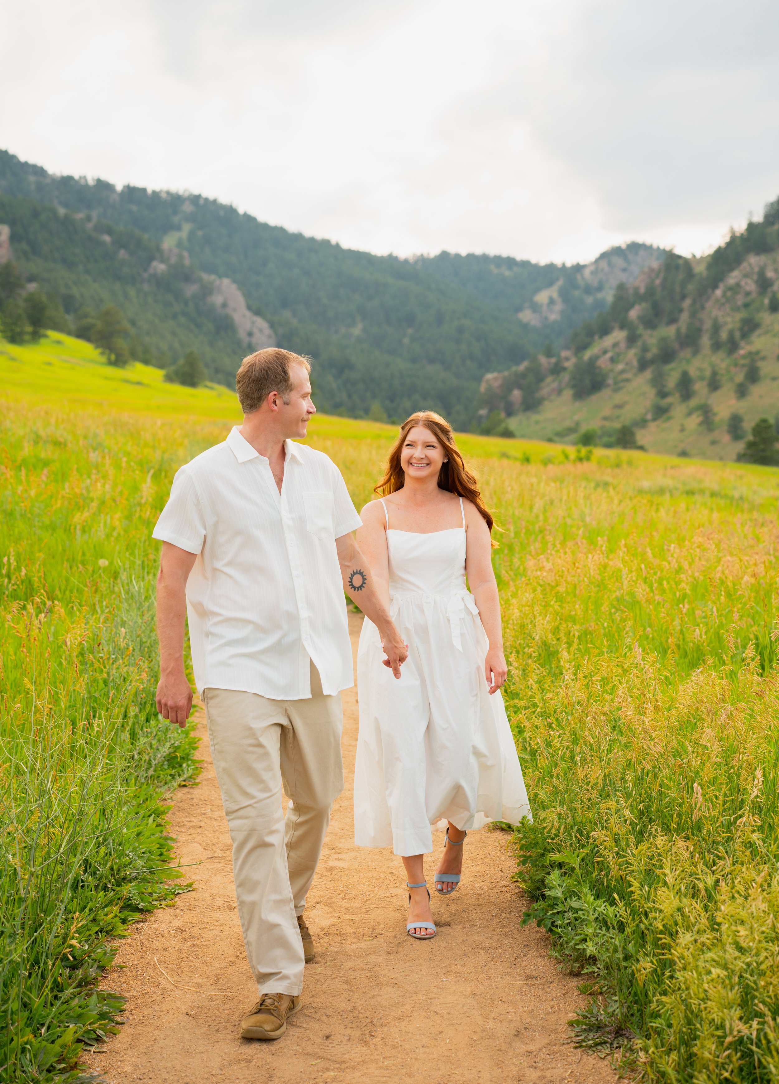 A happy couple walking on a dirt path through a green field with mountains in the background.