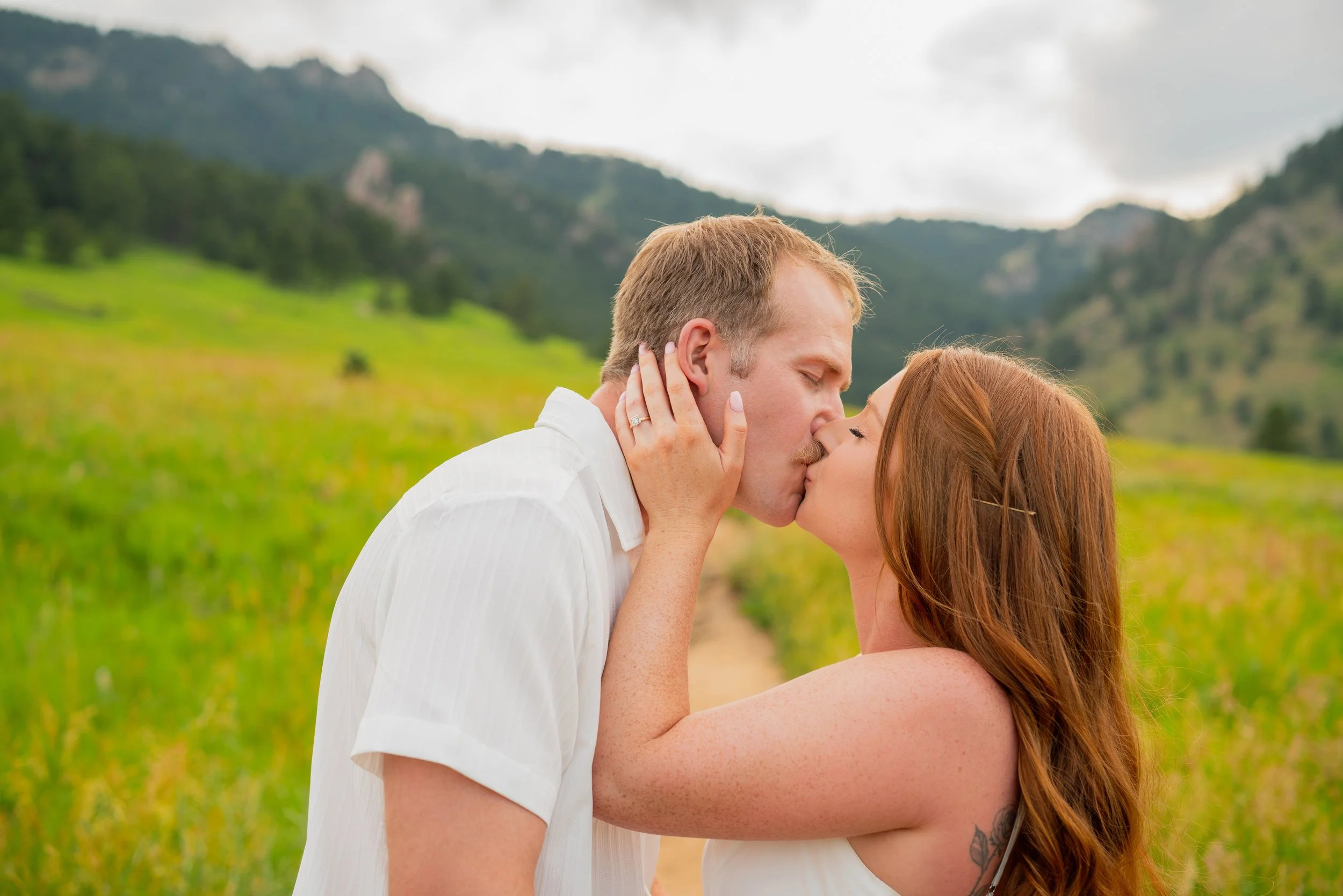 A couple shares a kiss outdoors in a green field with mountains in the background.