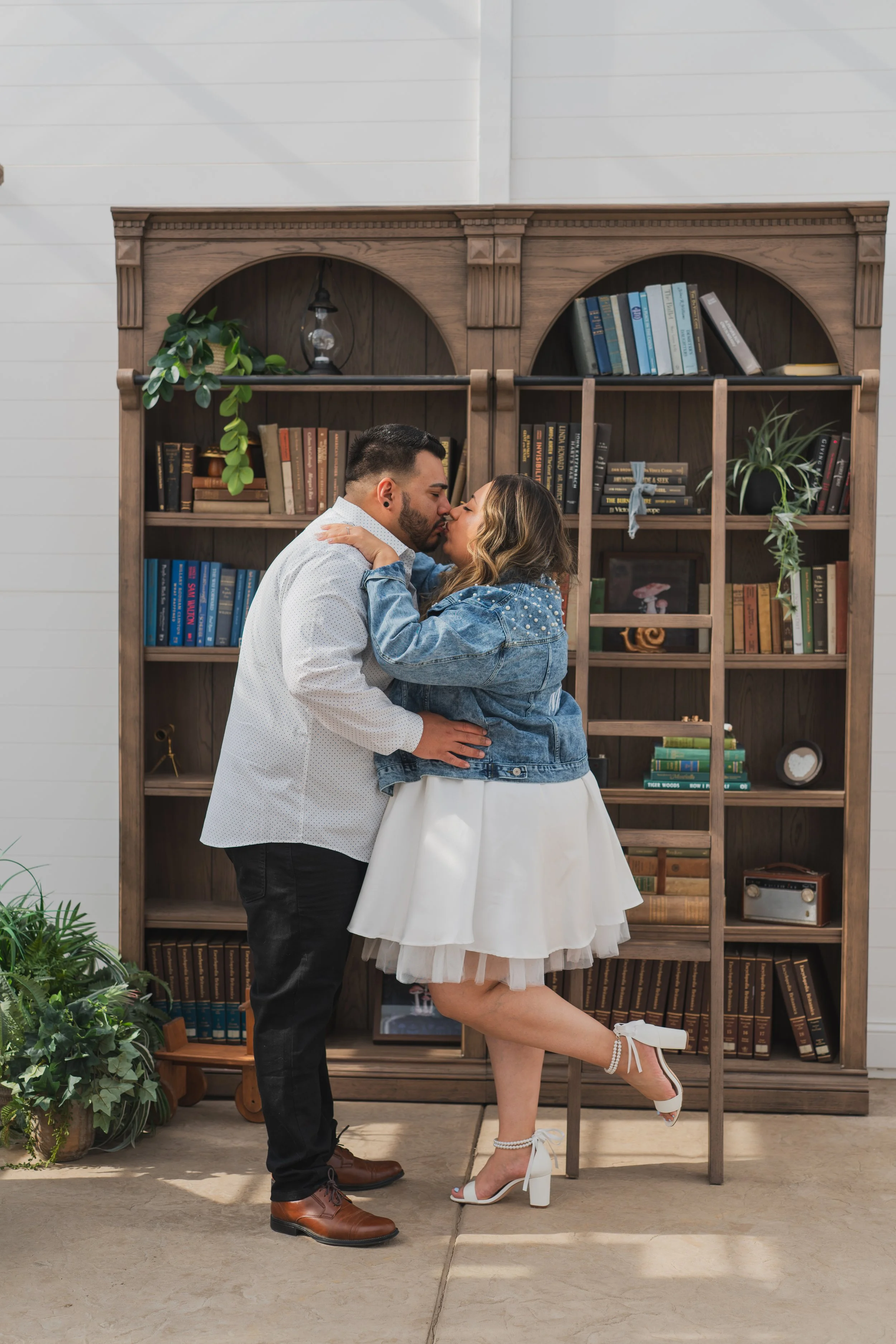 A couple is kissing in front of a wooden bookshelf. The man is wearing a white shirt and black pants, and the woman is wearing a denim jacket, white skirt, and white high heels. They are holding each other and standing on a concrete floor with a plan
