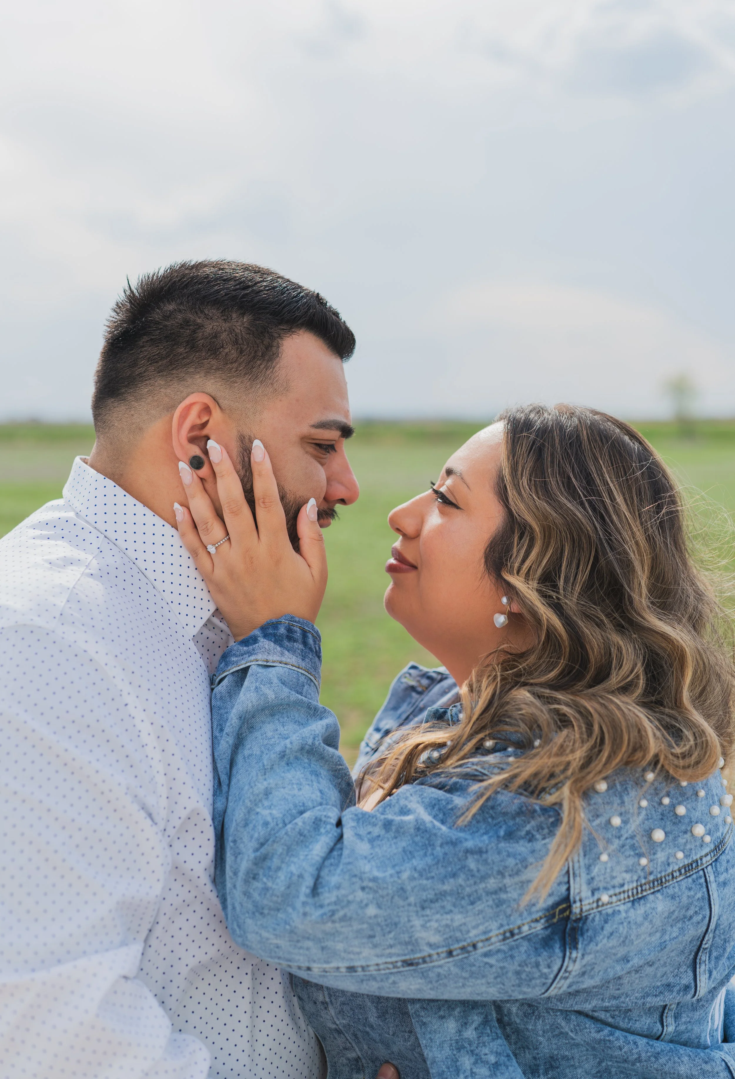 A couple facing each other outdoors, with the woman touching the man's face, both with closed eyes and gentle expressions.