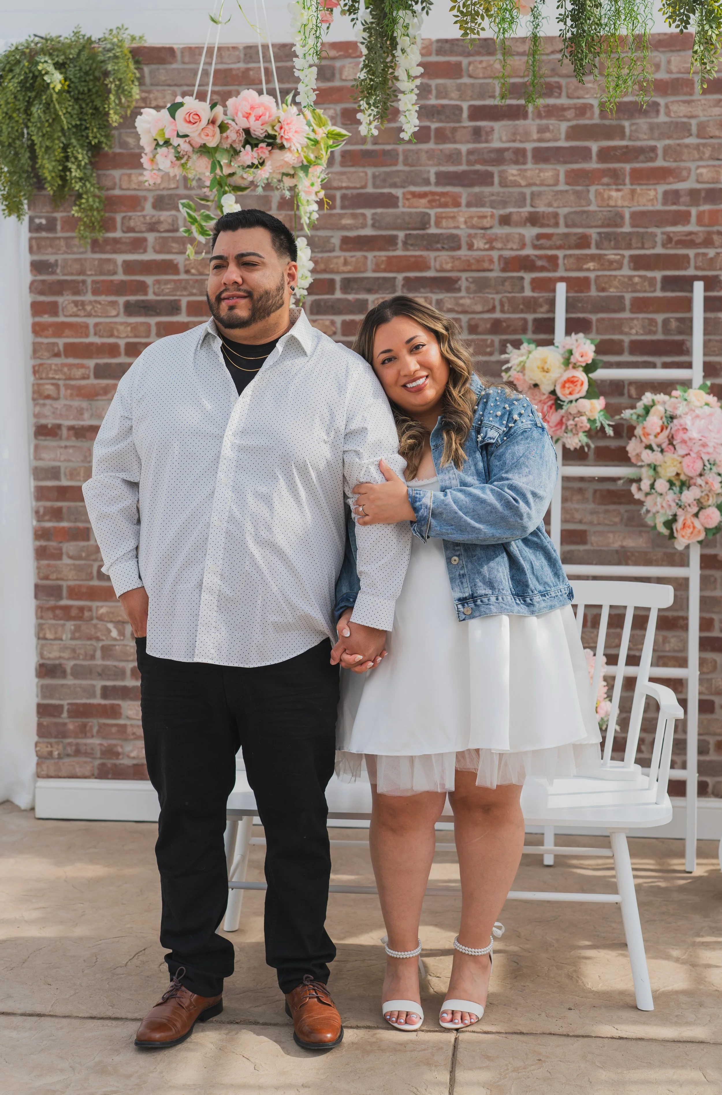 A smiling woman hugging a man in an indoor setting decorated with pink and white flowers, a white wooden bench, and a brick wall background.