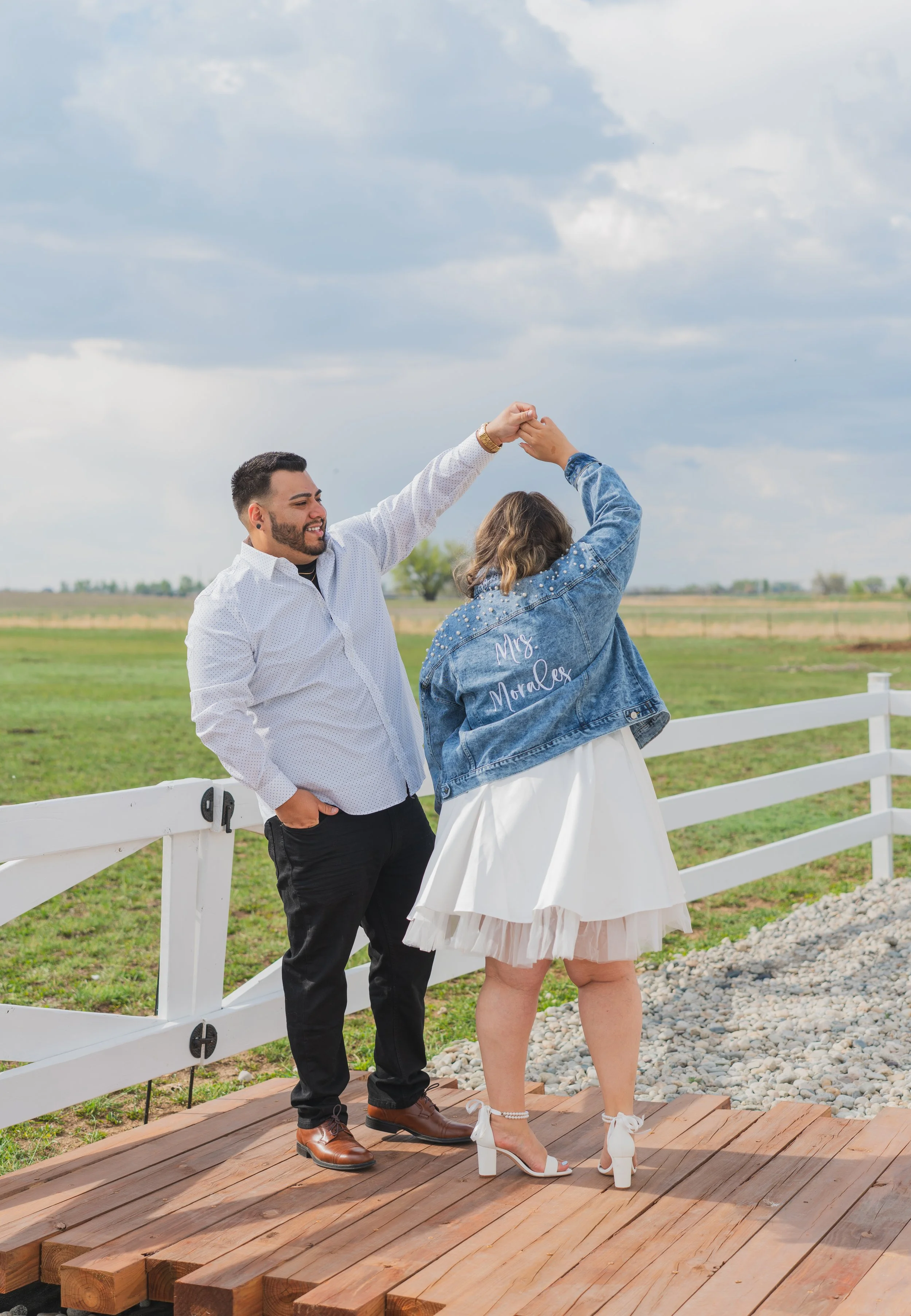 A couple, a man and a woman, dancing on on a wooden deck outdoors. The woman is wearing a white dress, high heels, and a denim jacket with 