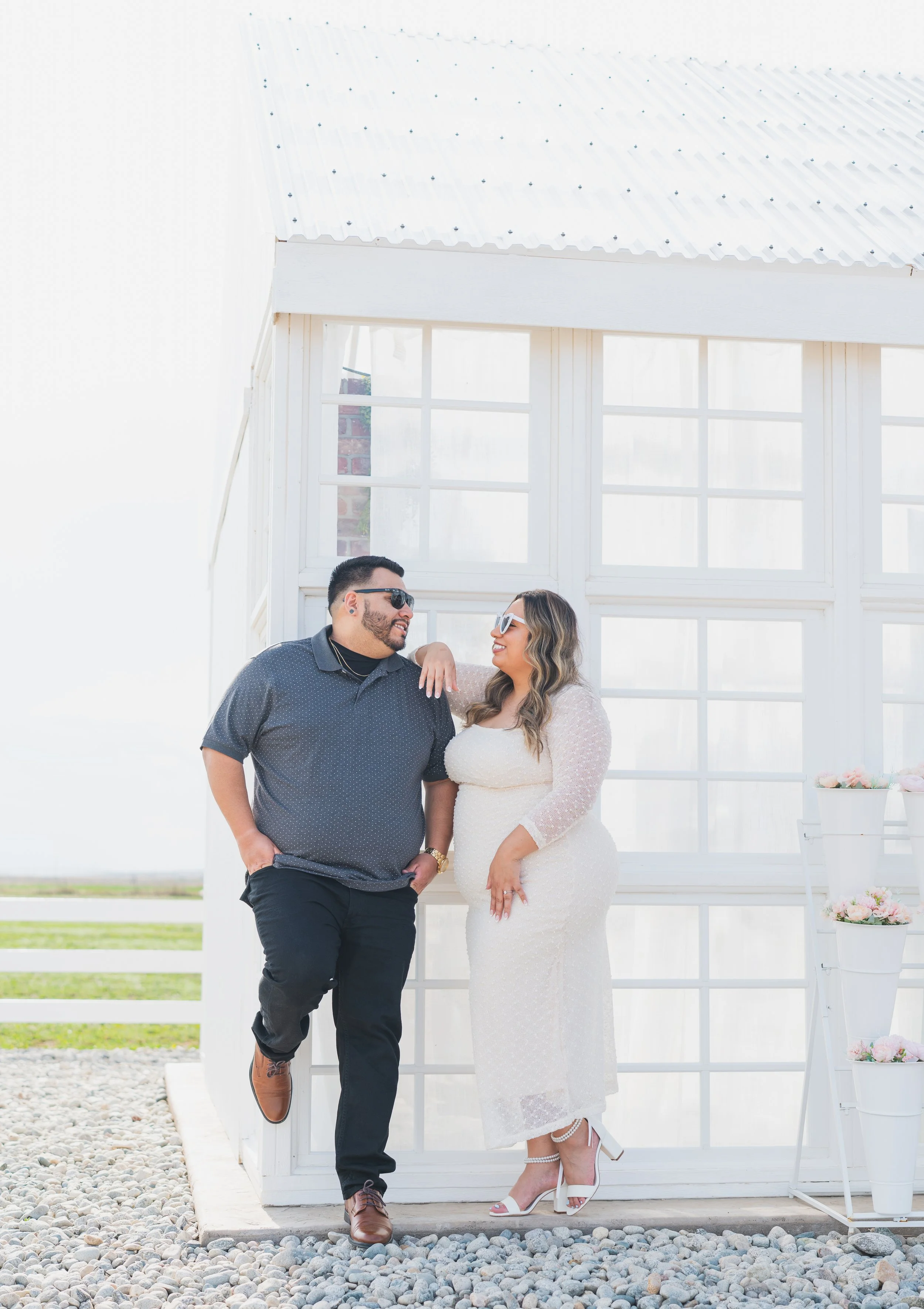 A couple dressed in wedding attire standing outside in front of a white building with large windows, smiling and looking at each other.