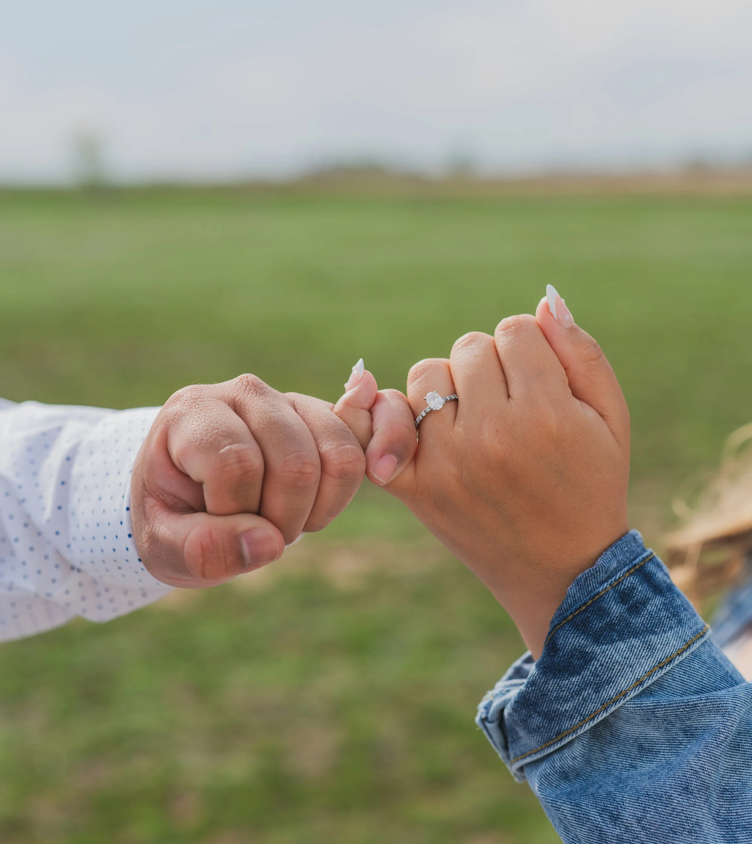 Close-up of a man and woman holding hands with the woman's engagement ring visible, outdoors in a grassy field.
