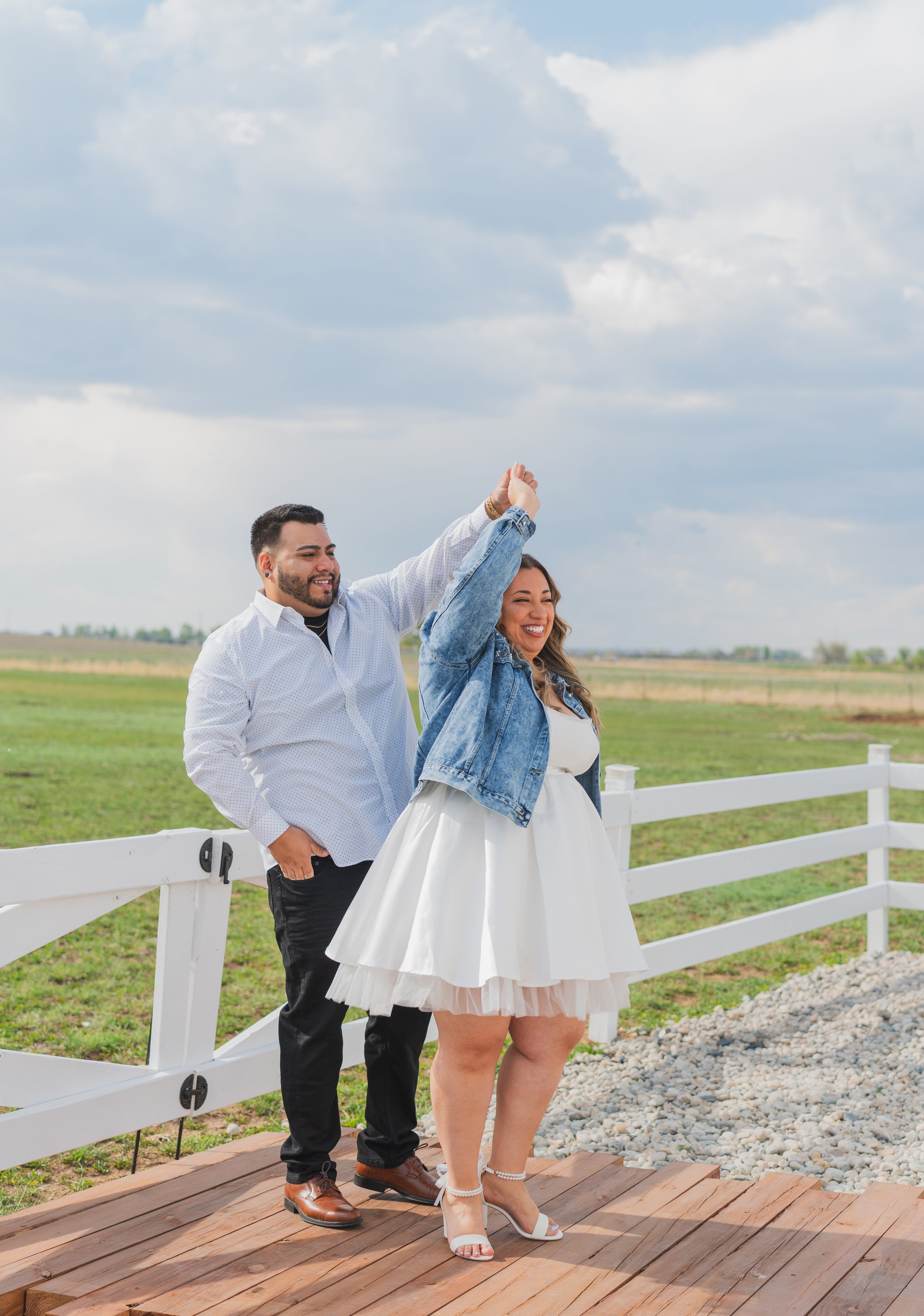 A couple dancing outdoors on a wooden platform, with the man holding the woman's hand and both smiling, in a rural setting with a white fence and open fields under a cloudy sky.