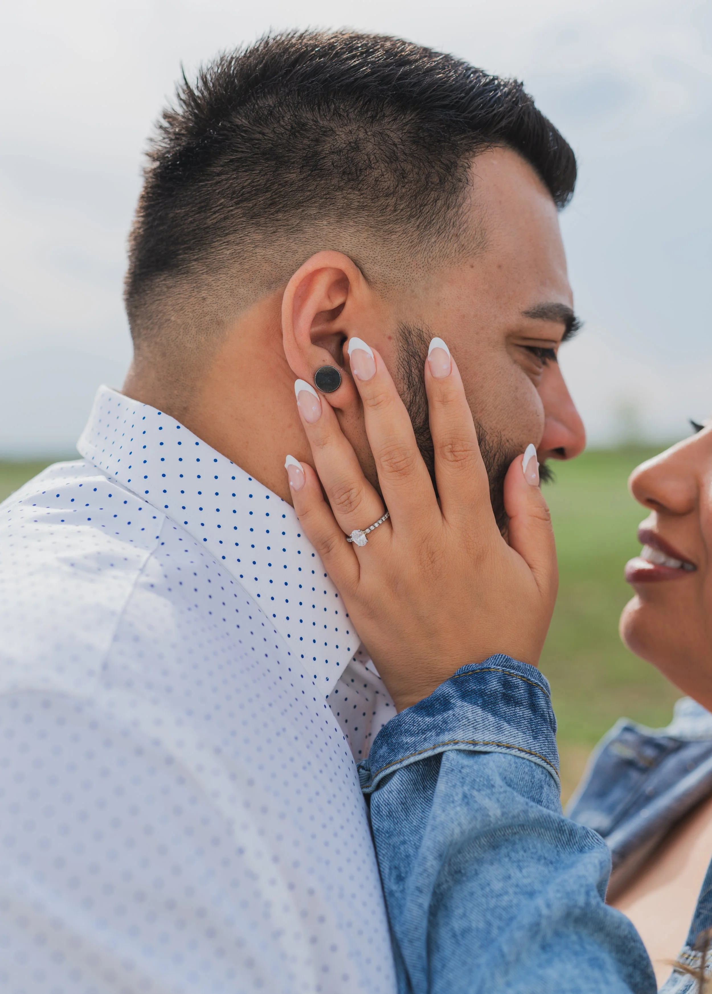 A couple is close, with the woman holding the man's face and showing a diamond engagement ring on her finger. The man is smiling with eyes closed, wearing a white shirt with blue polka dots, with short dark hair and a beard. They are outdoors.