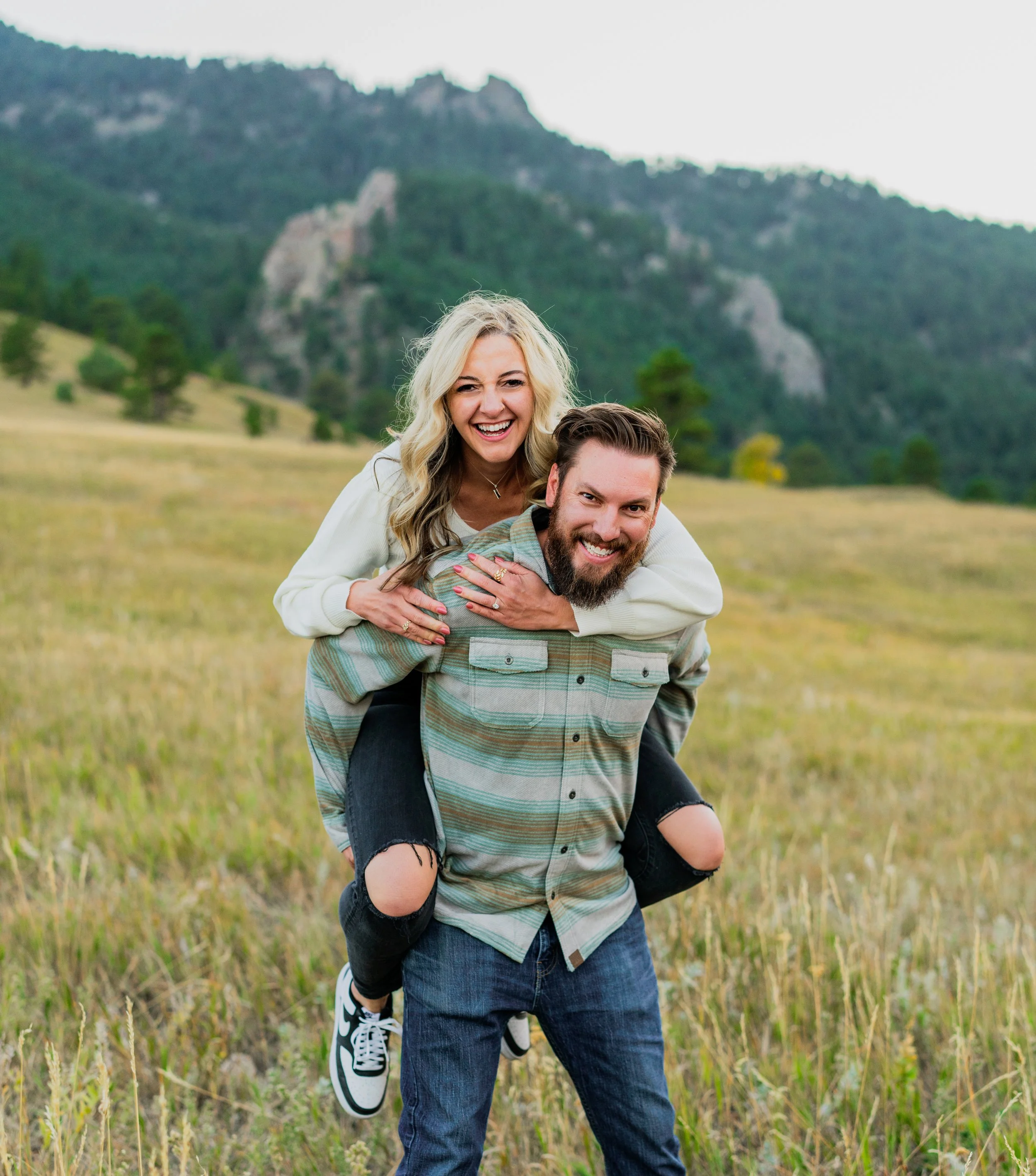 A smiling couple enjoying outdoor activities in a grassy field with a mountain background. The woman is on the man's back, and they are both smiling.