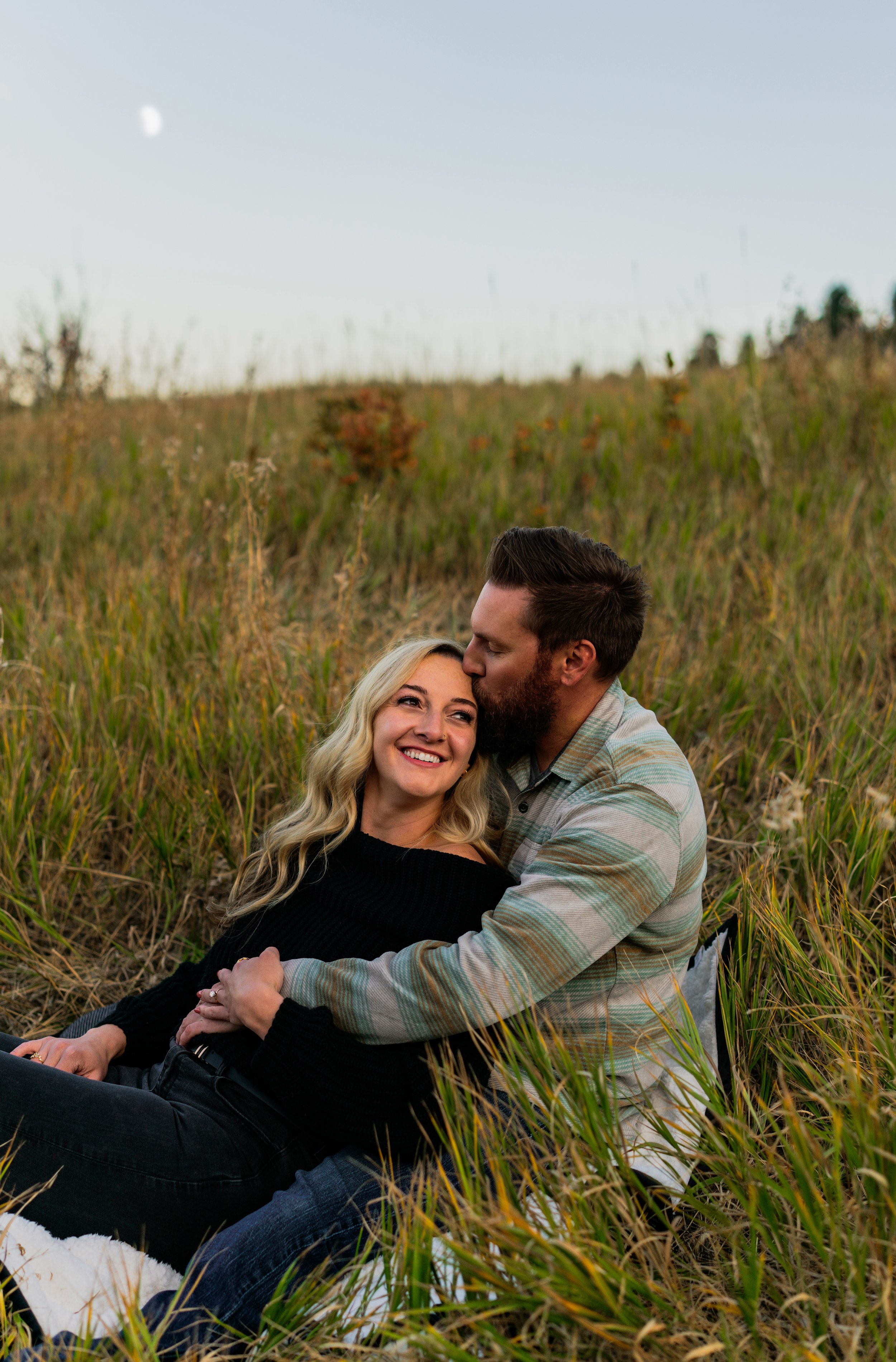A couple sitting in a grassy field, smiling and kissing, during sunset with the moon visible in the sky.