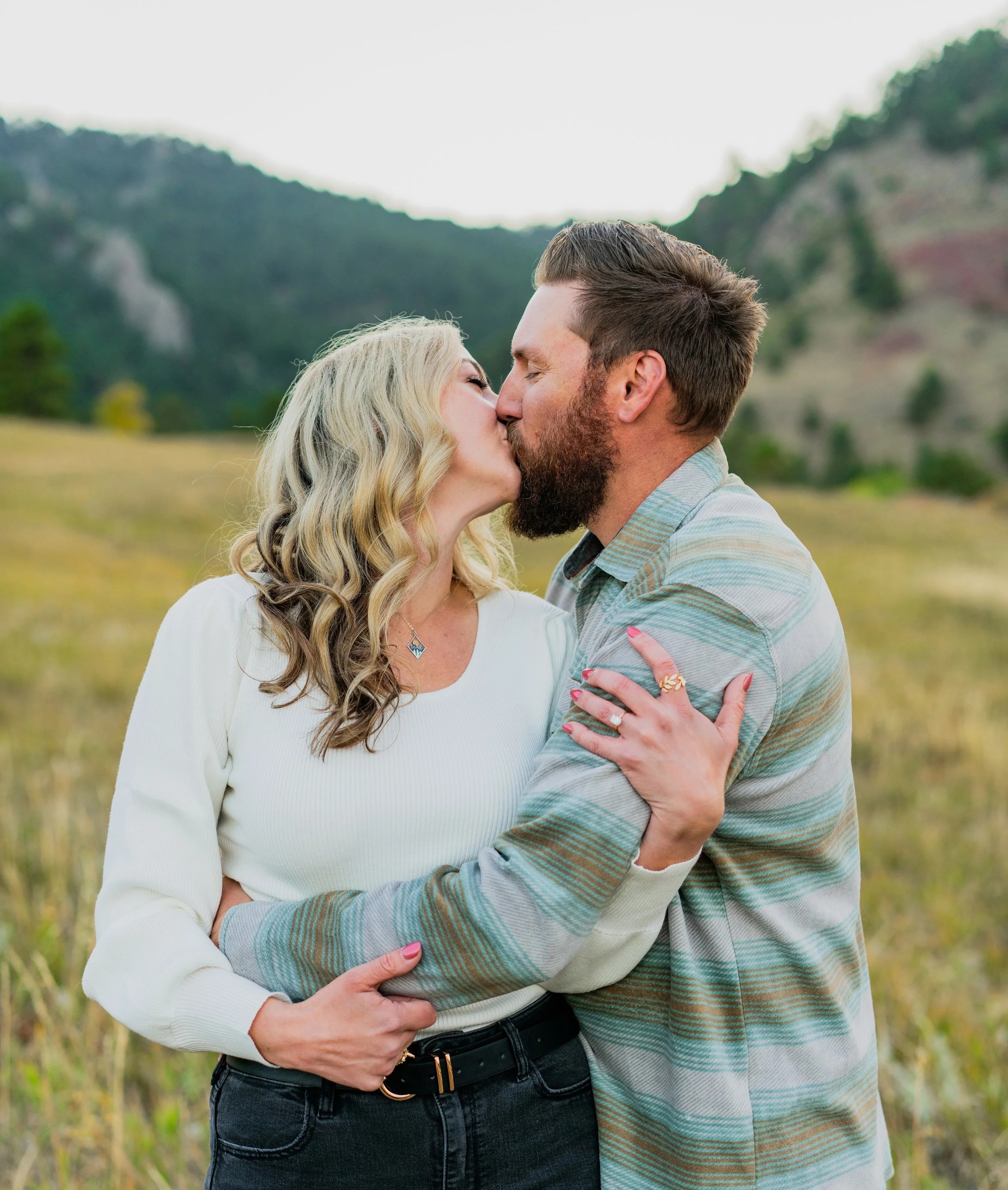 A couple kissing in a field with mountains and trees in the background.