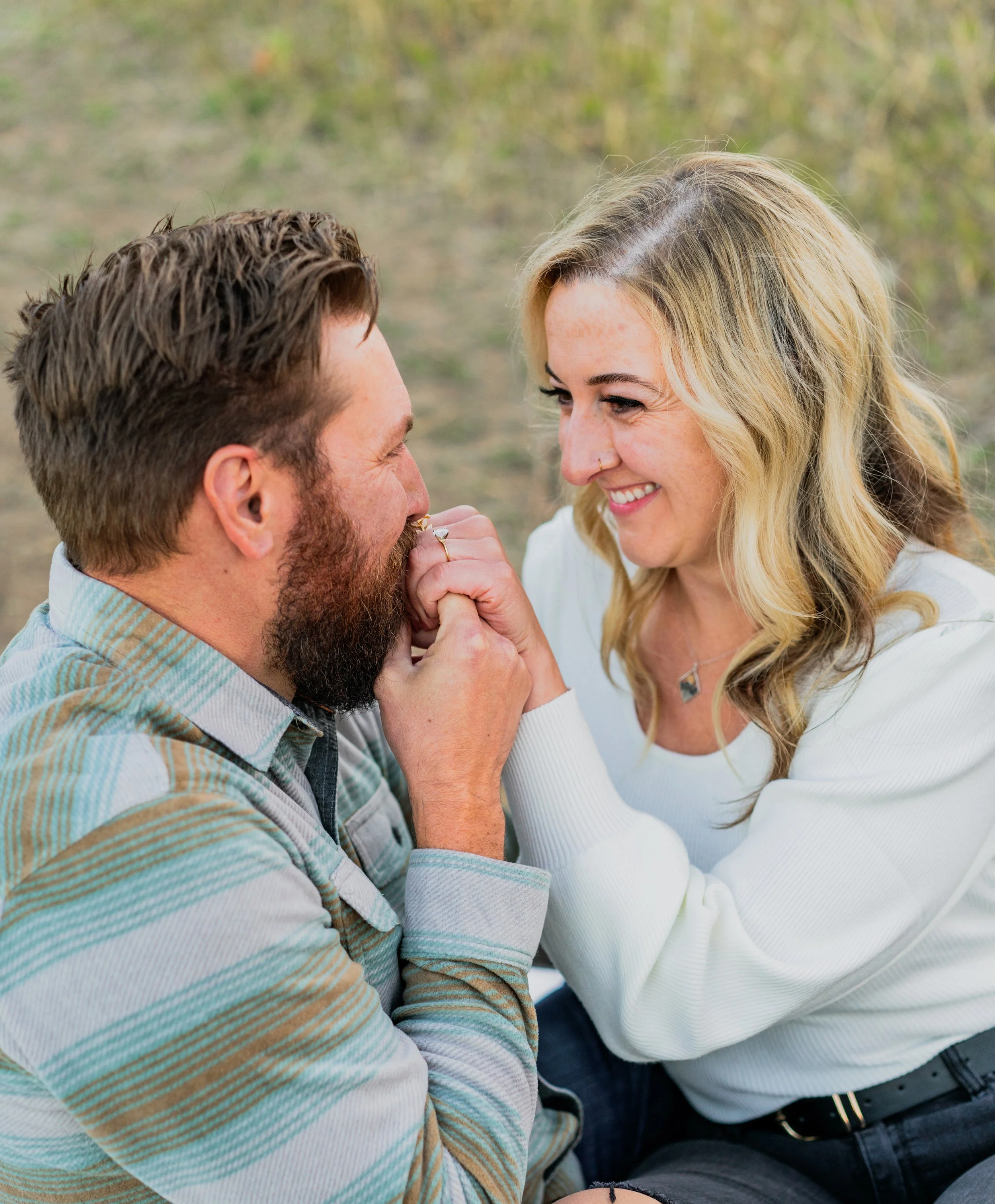 A man with a beard and a woman with blonde hair smiling at each other, holding hands near their faces outdoors.