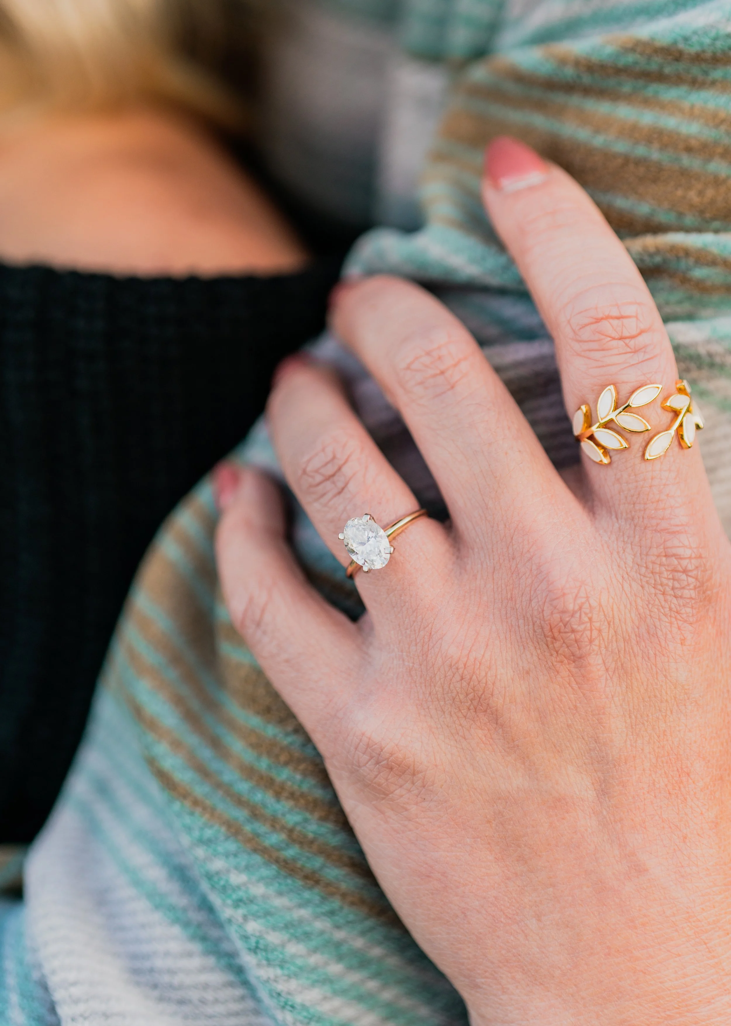 Close-up of a hand displaying a ring with a large gemstone and a gold ring with leaf-shaped design, resting on a striped fabric surface.