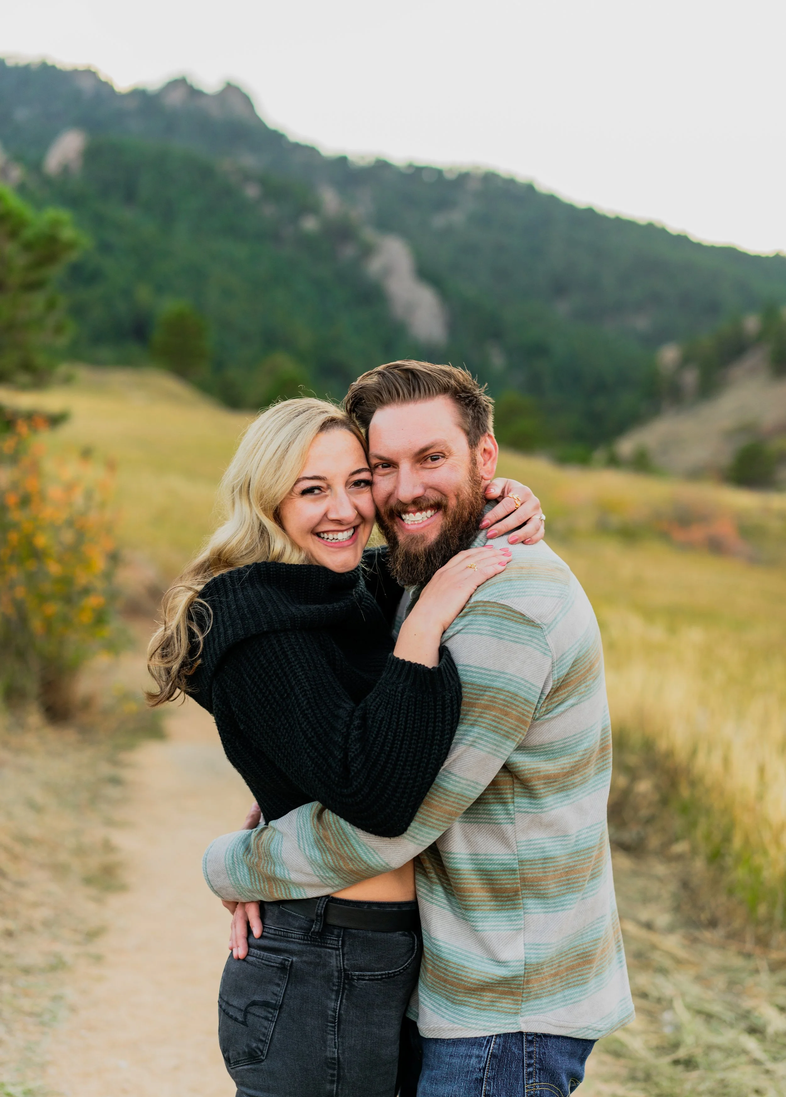 A happy couple hugging outdoors in a scenic mountain landscape with green hills and cloudy sky.
