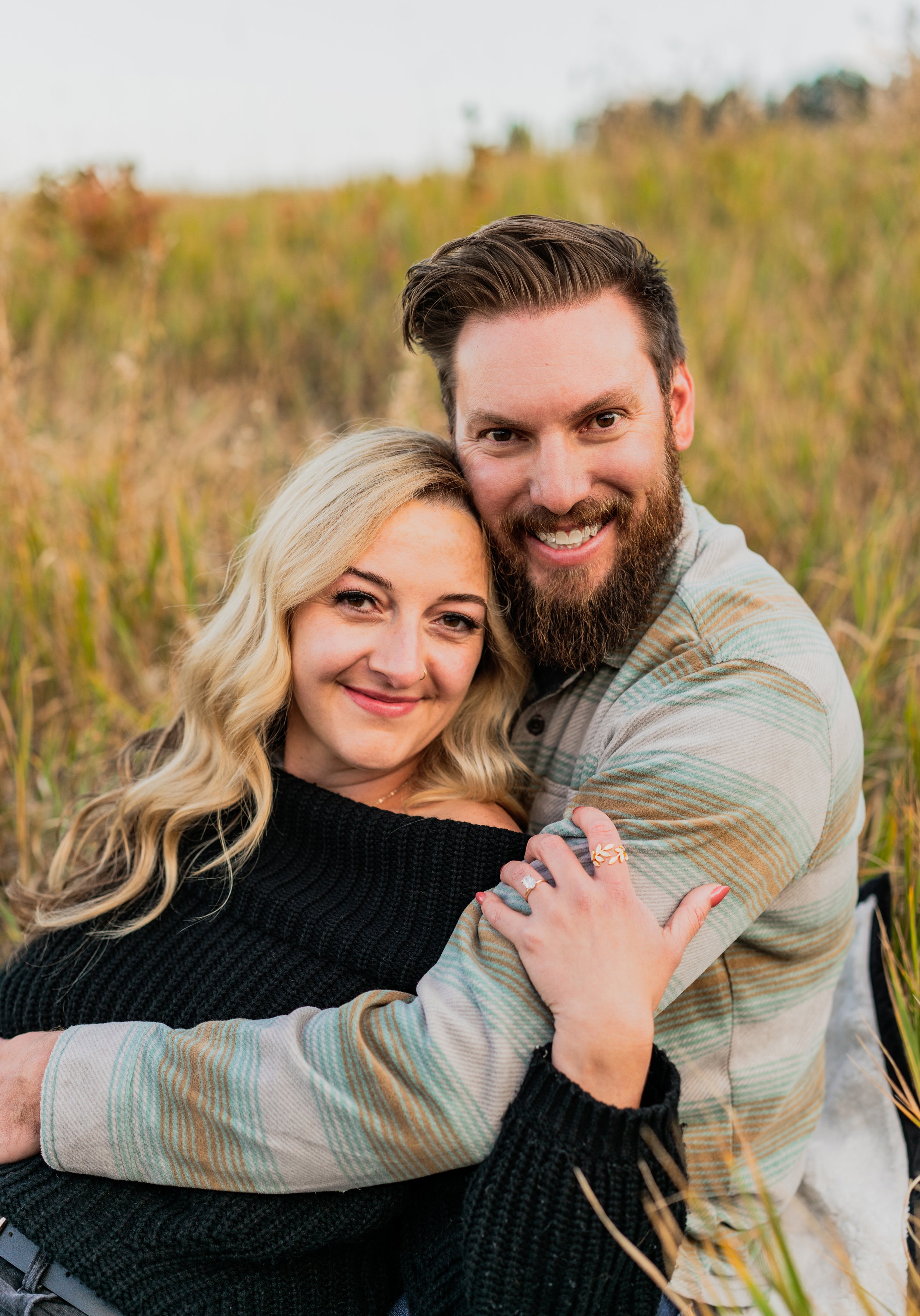 A smiling couple embracing outdoors in a field of tall grass, with a blurred background of trees.