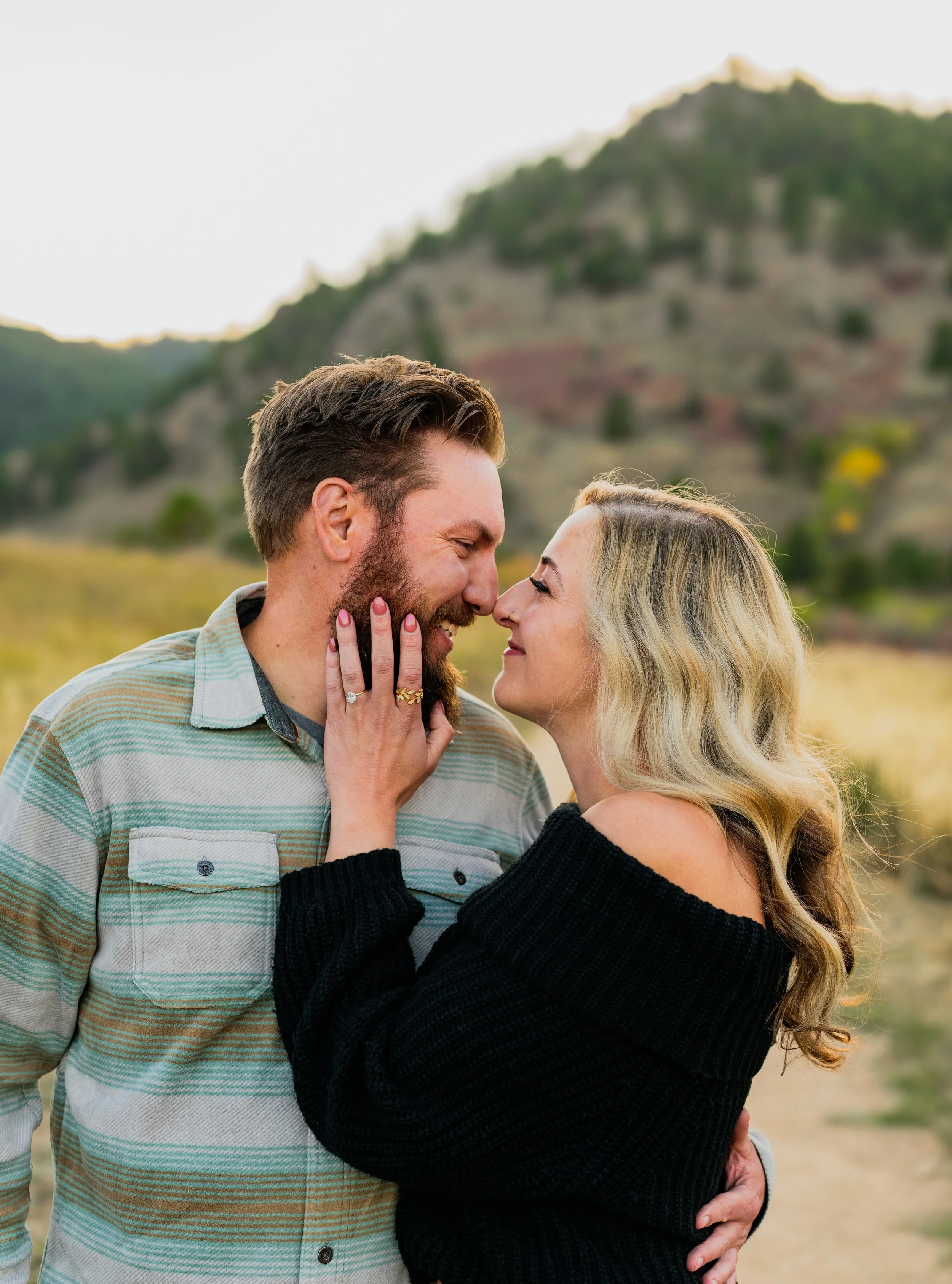 A couple face to face in an outdoor setting with mountains in the background, smiling and touching their noses together.