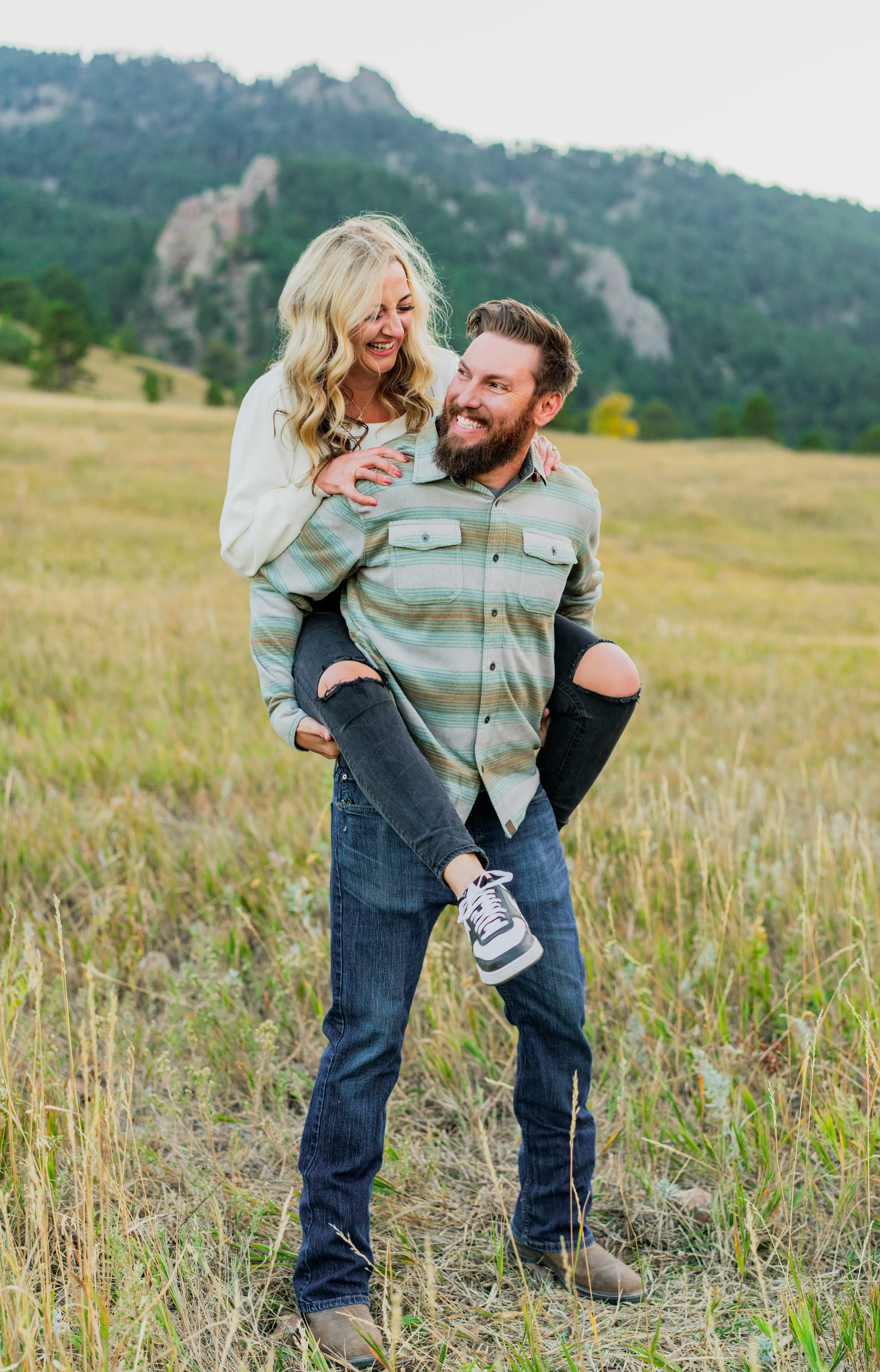 A happy couple enjoying a walk in a grassy field with mountains and trees in the background. The woman is riding on the man's back, both smiling and looking at each other.