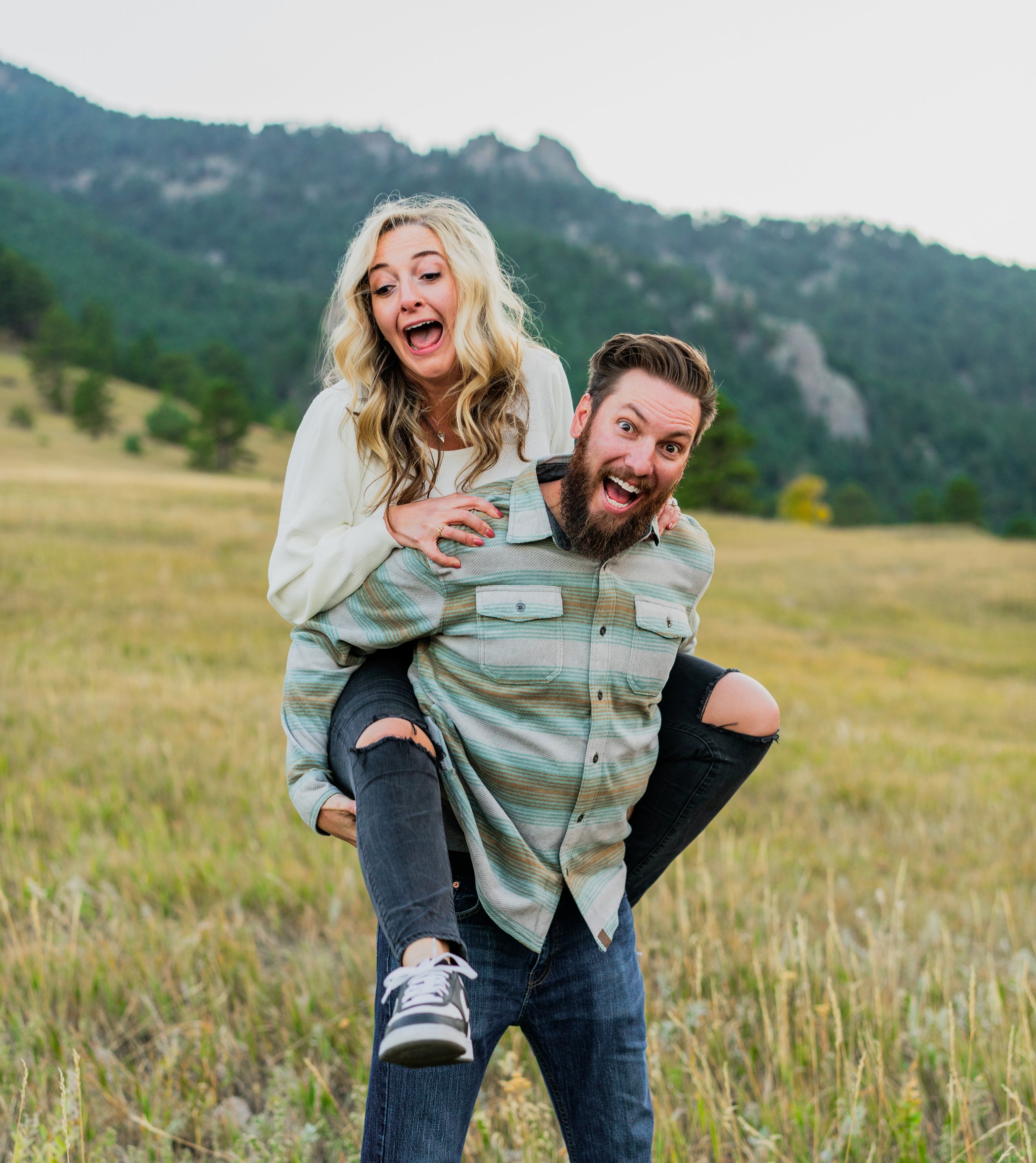 A couple enjoying a piggyback ride outdoors in a grassy field with mountains in the background.