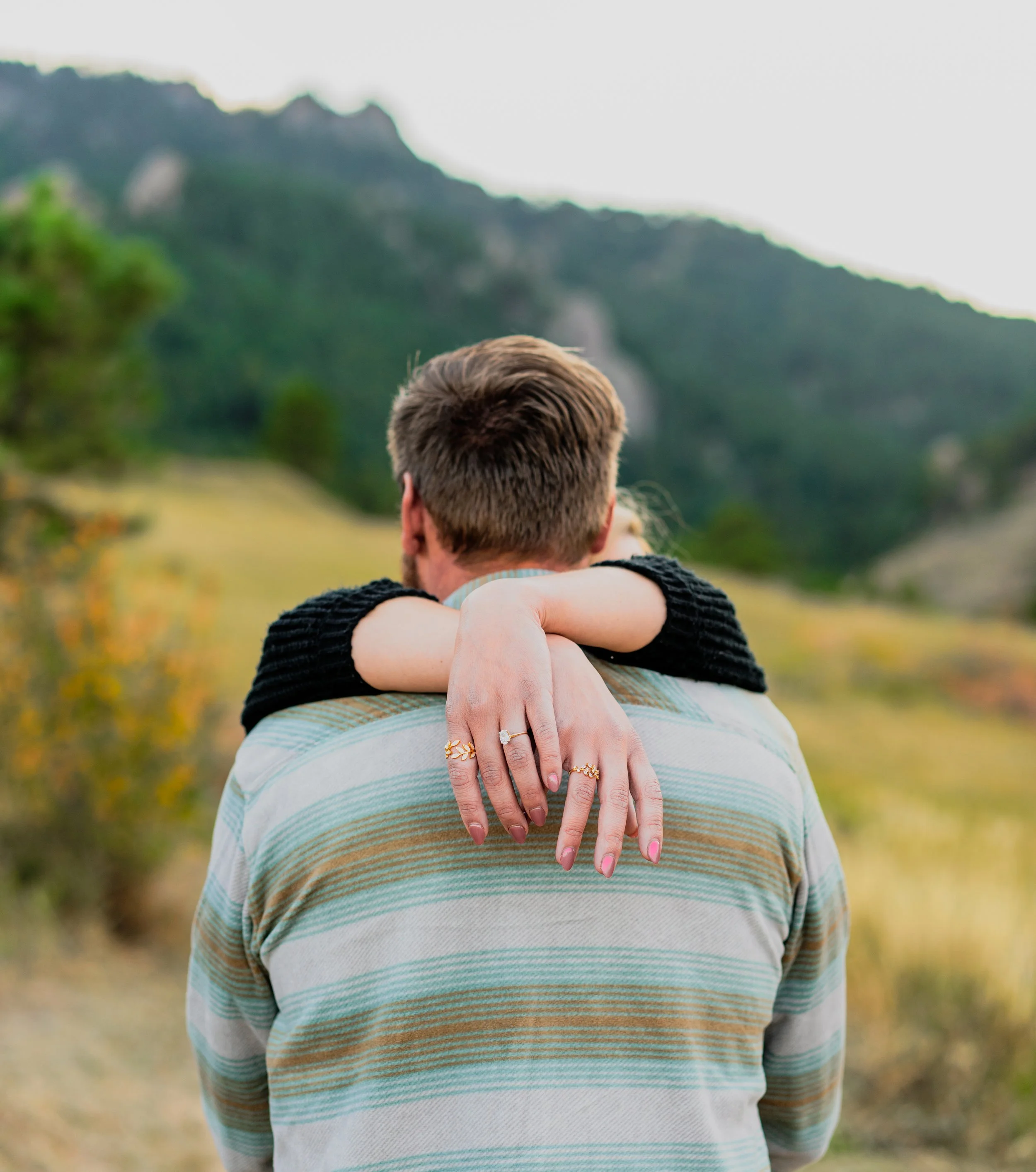A man with short brown hair and a striped shirt is seen from behind, with a woman's arms around his neck, showing her hands with rings, in an outdoor natural setting with mountains and greenery in the background.