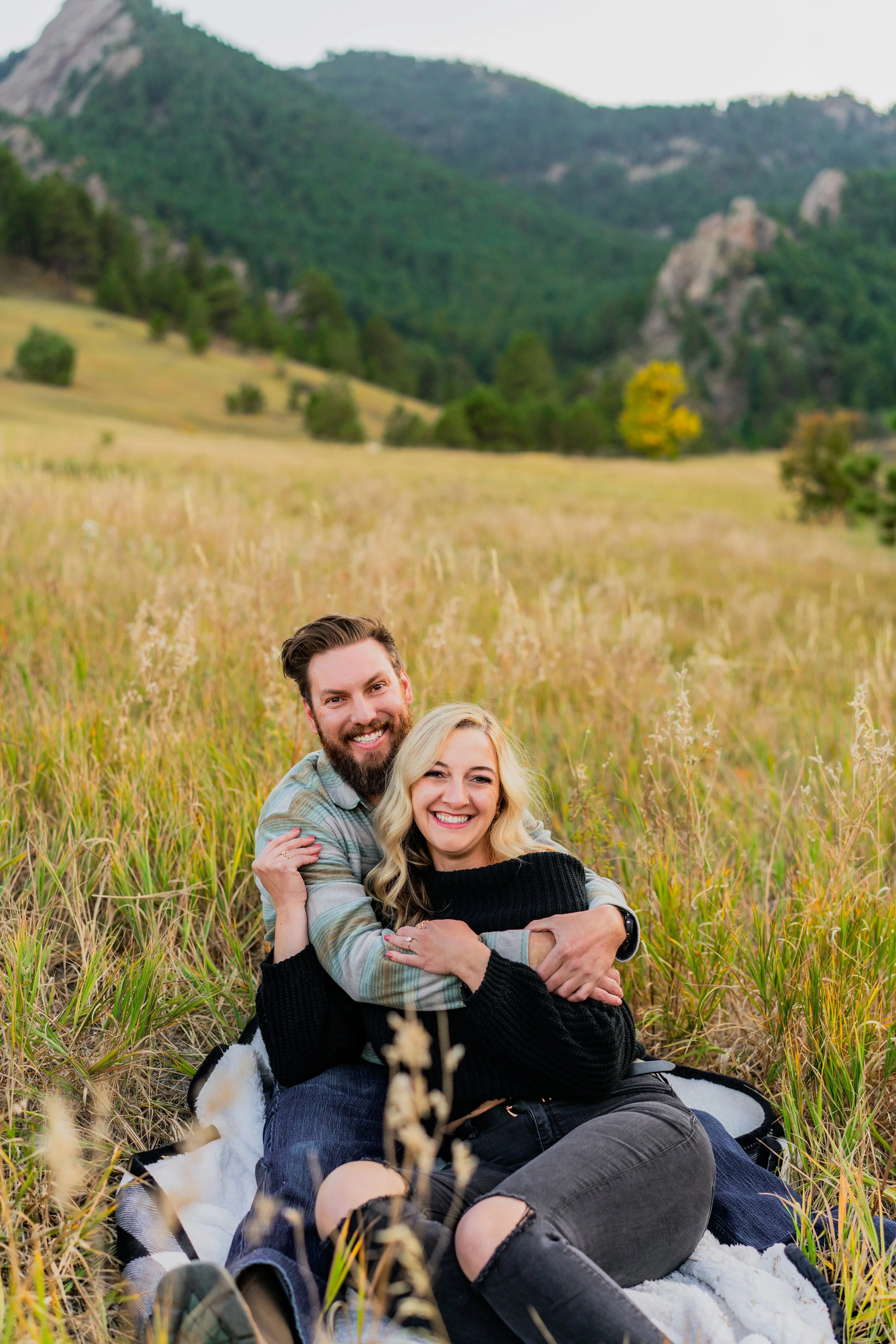 A happy couple sitting on a blanket in a grassy field with mountains in the background.