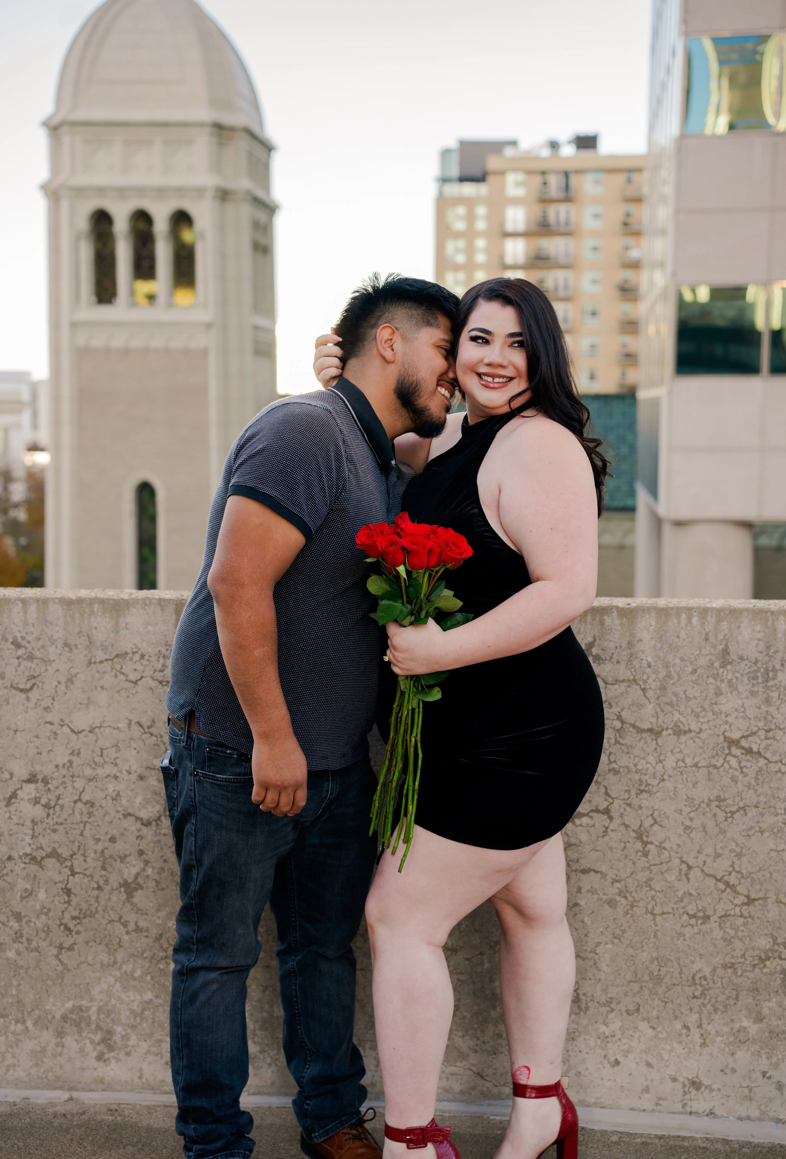 A couple standing on a rooftop, with a cityscape and a church tower in the background, smiling, with the woman holding a bouquet of red roses.