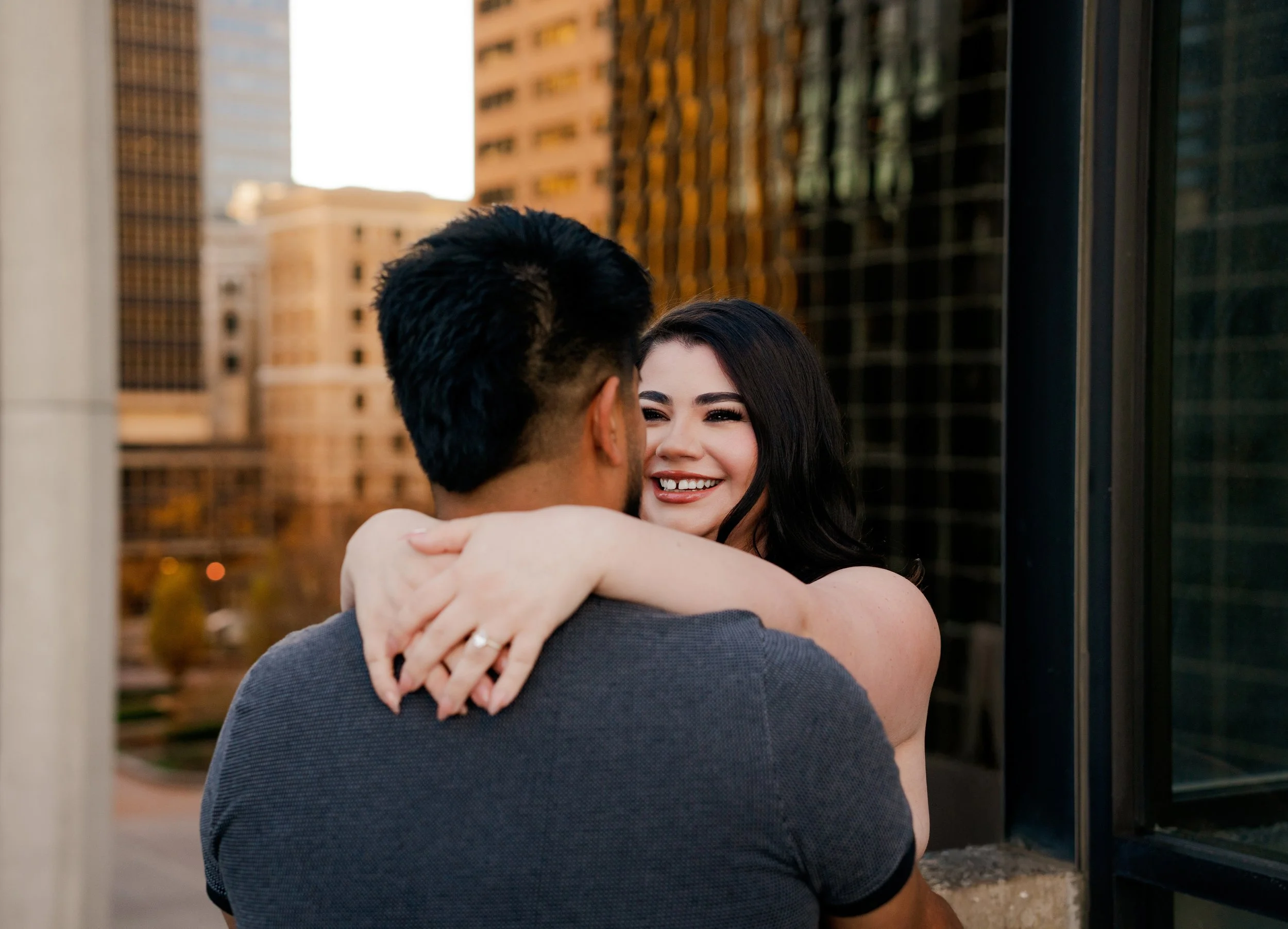 A woman with black hair and a big smile hugging a man with dark hair in an urban setting during sunset.